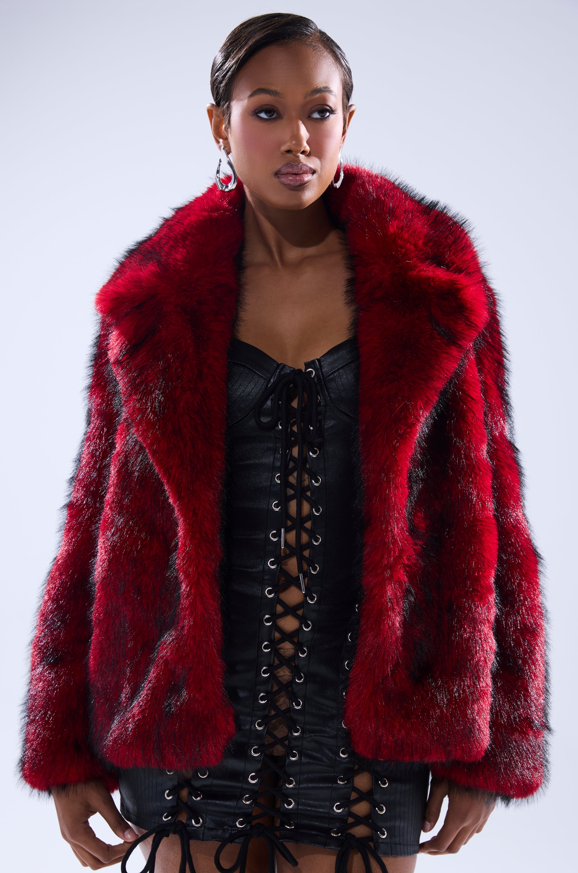 Wearing the NO LOOKING BACK FAUX FUR IN RED MULTI over a black lace-up dress, a woman stands against a light background, gazing to the side with confidence.