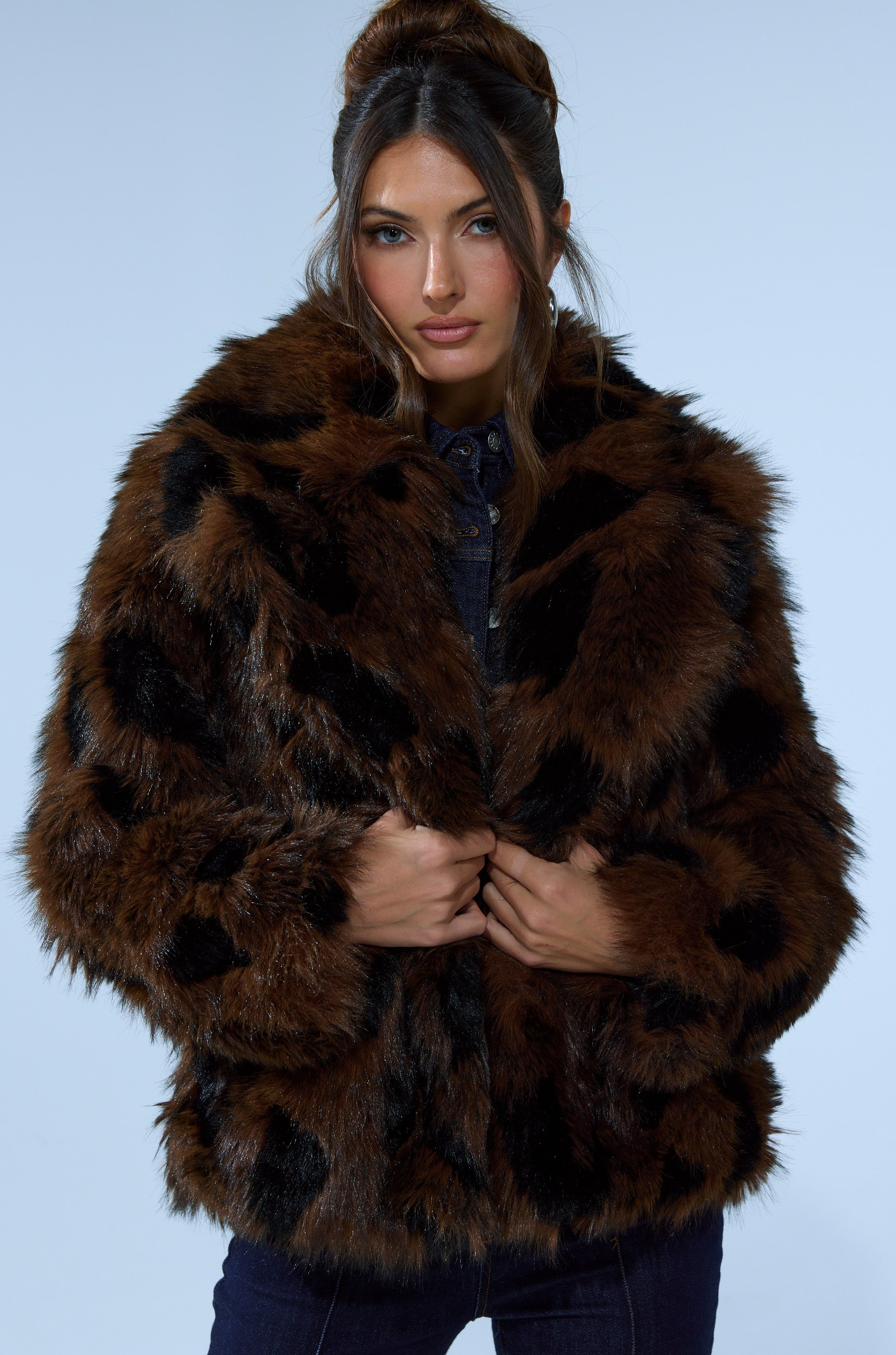 A woman with long brown hair styled in an updo wears the SHORTIE FUR COAT IN BROWN MULTI over a dark outfit, standing against a light blue background. She faces the camera, holding the coat closed.