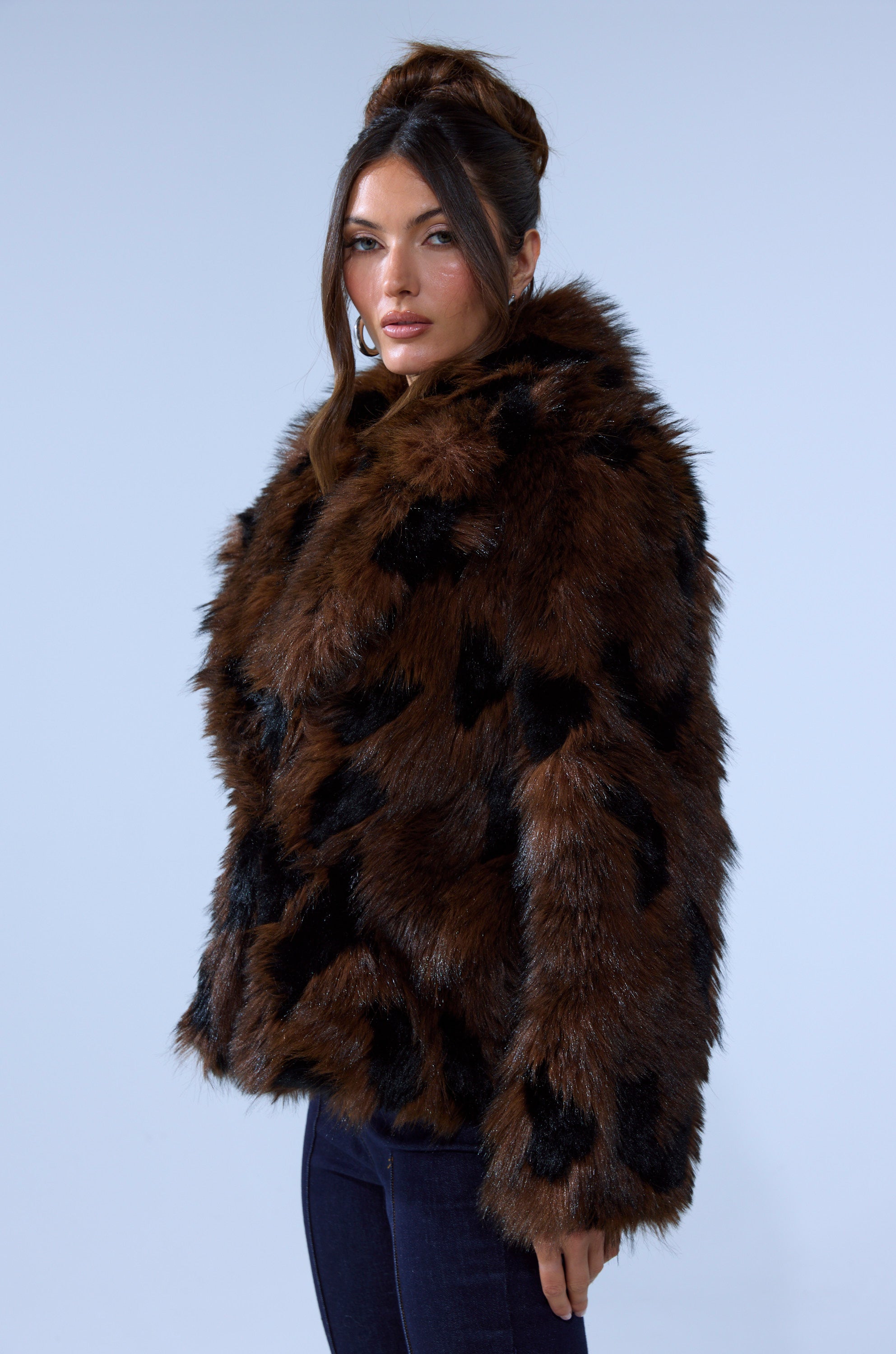 A woman with long brown hair styled in an updo models the SHORTIE FUR COAT IN BROWN MULTI and dark pants, standing against a plain light blue background.