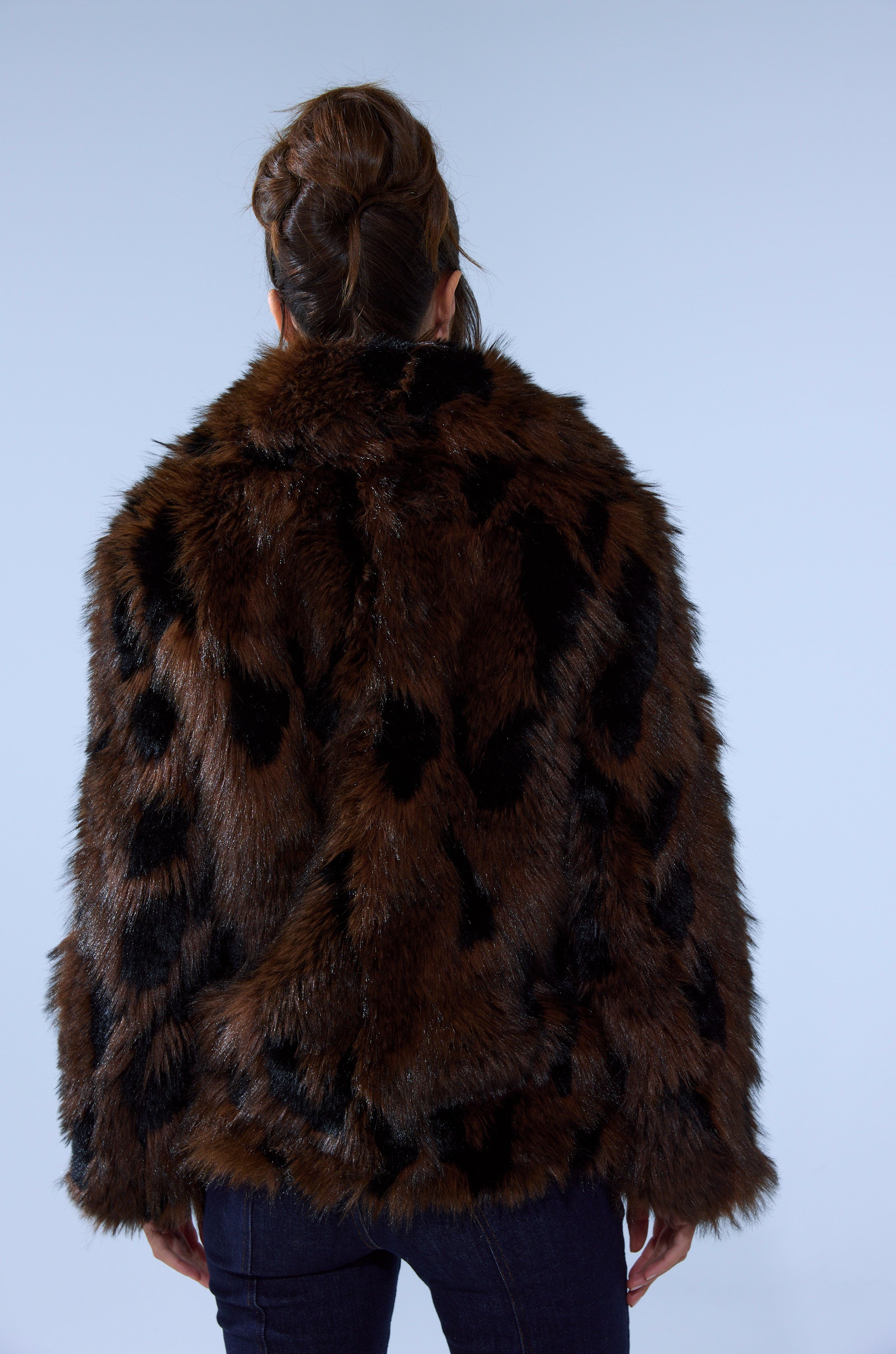 A person with brown hair in an updo is seen from behind, wearing the SHORTIE FUR COAT IN BROWN MULTI and dark jeans against a plain light background.