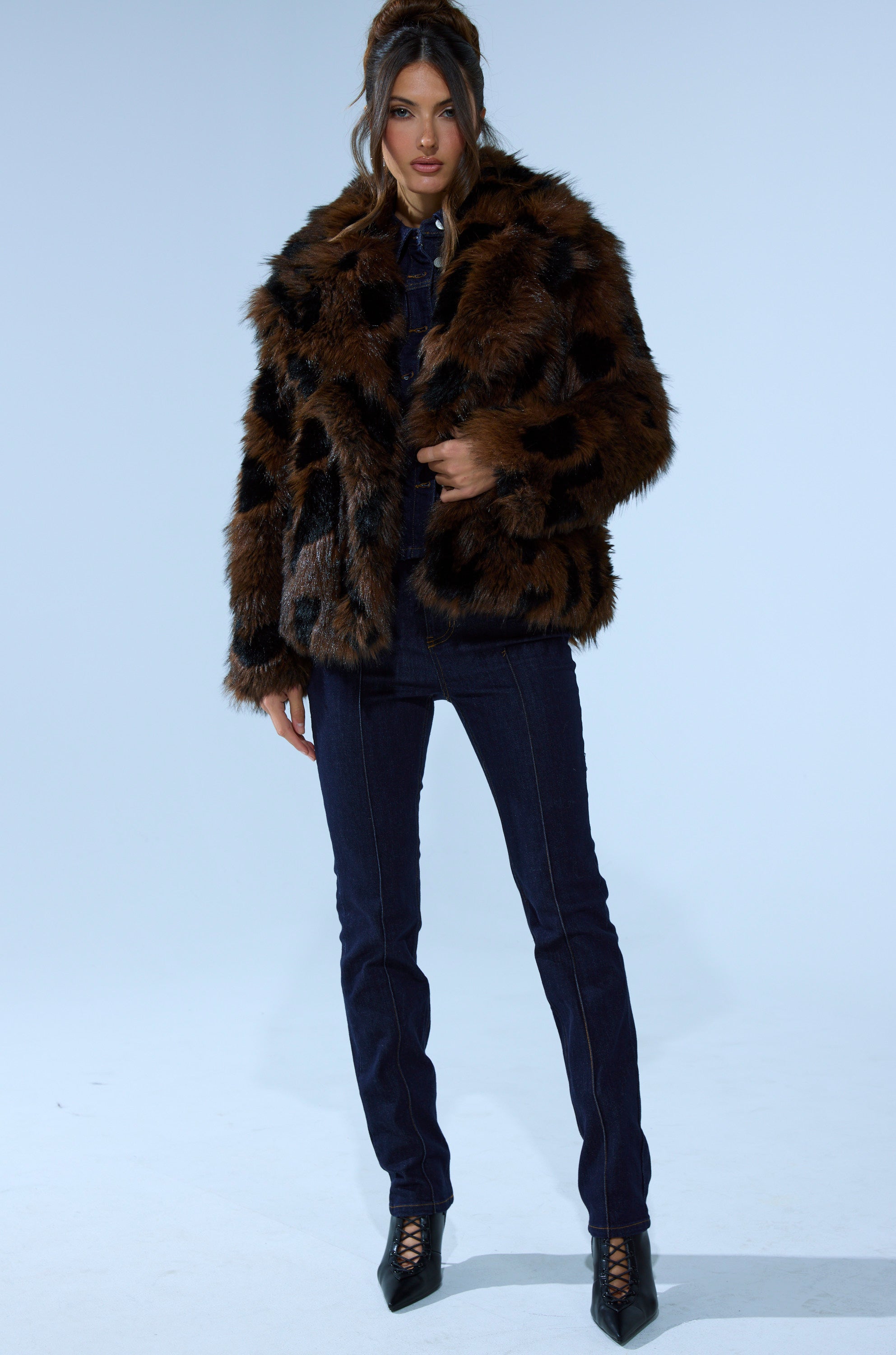 A woman stands confidently against a light blue background, wearing the SHORTIE FUR COAT IN BROWN MULTI, paired with dark denim jeans and black heeled lace-up boots.