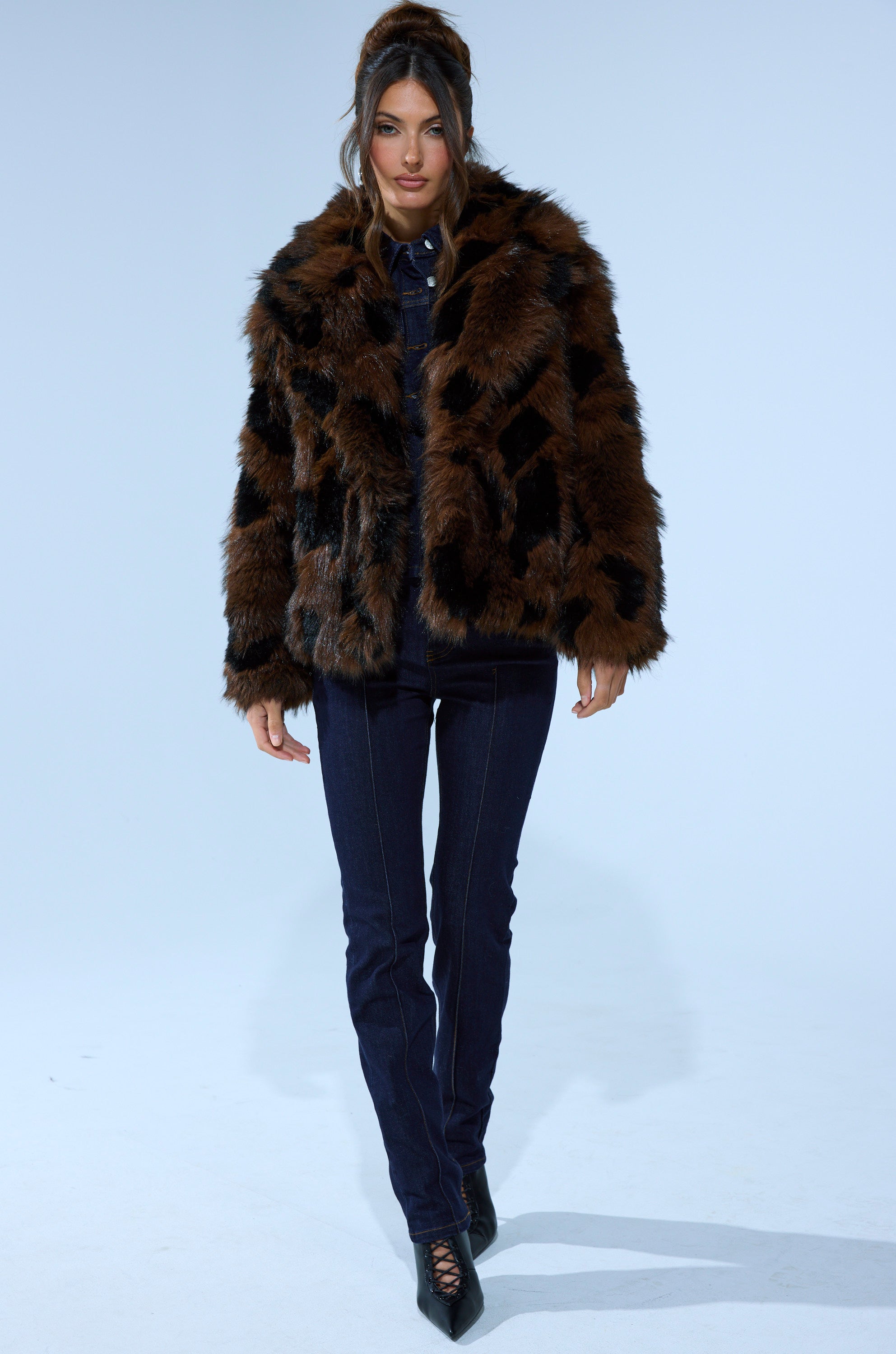 A woman with long, styled hair and a confident look wears the SHORTIE FUR COAT IN BROWN MULTI over dark denim and black pointed shoes, standing against a plain background.