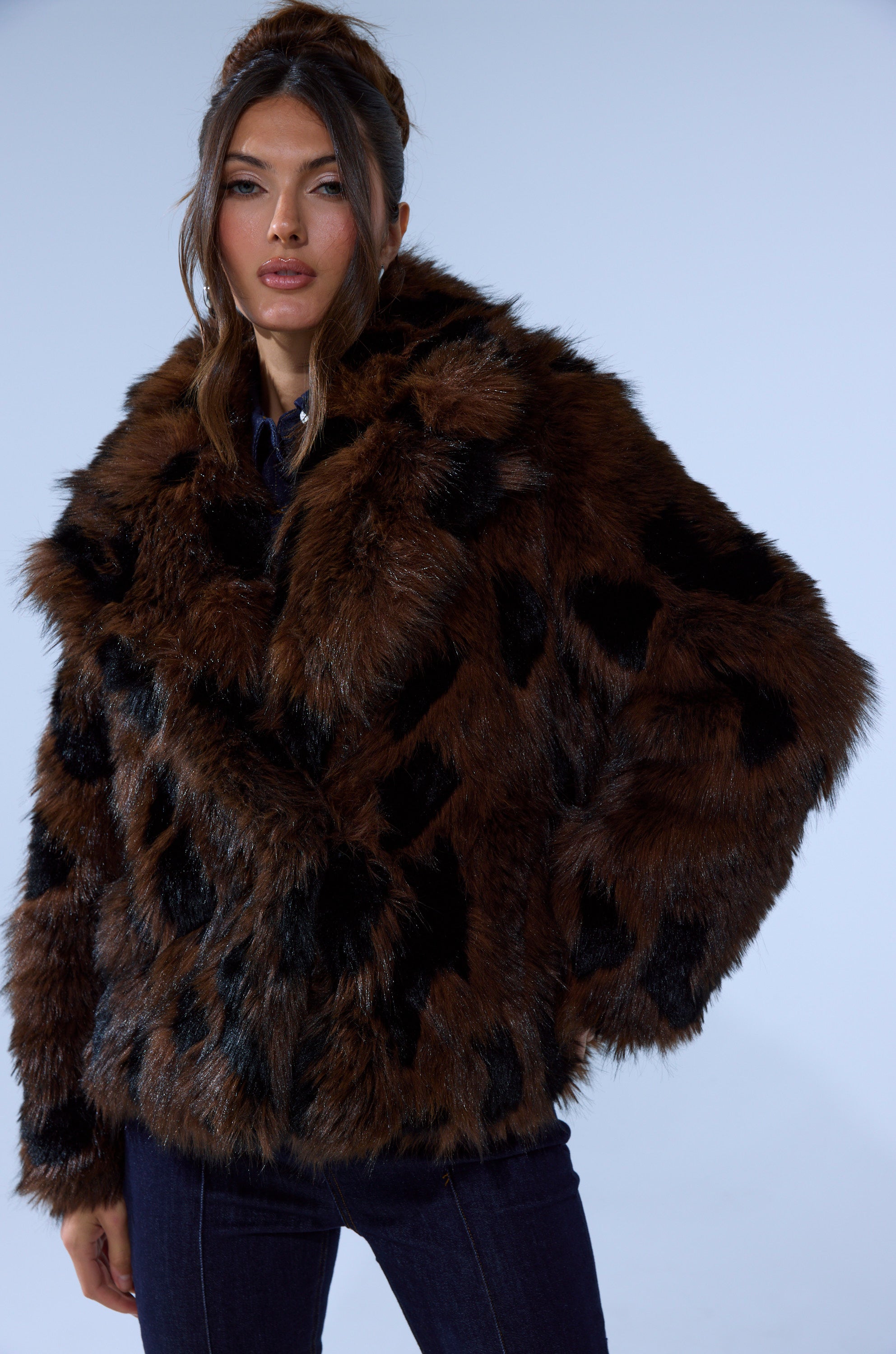 A woman with long brown hair models the SHORTIE FUR COAT IN BROWN MULTI, featuring oversized brown fur with black accents, against a plain light backdrop.