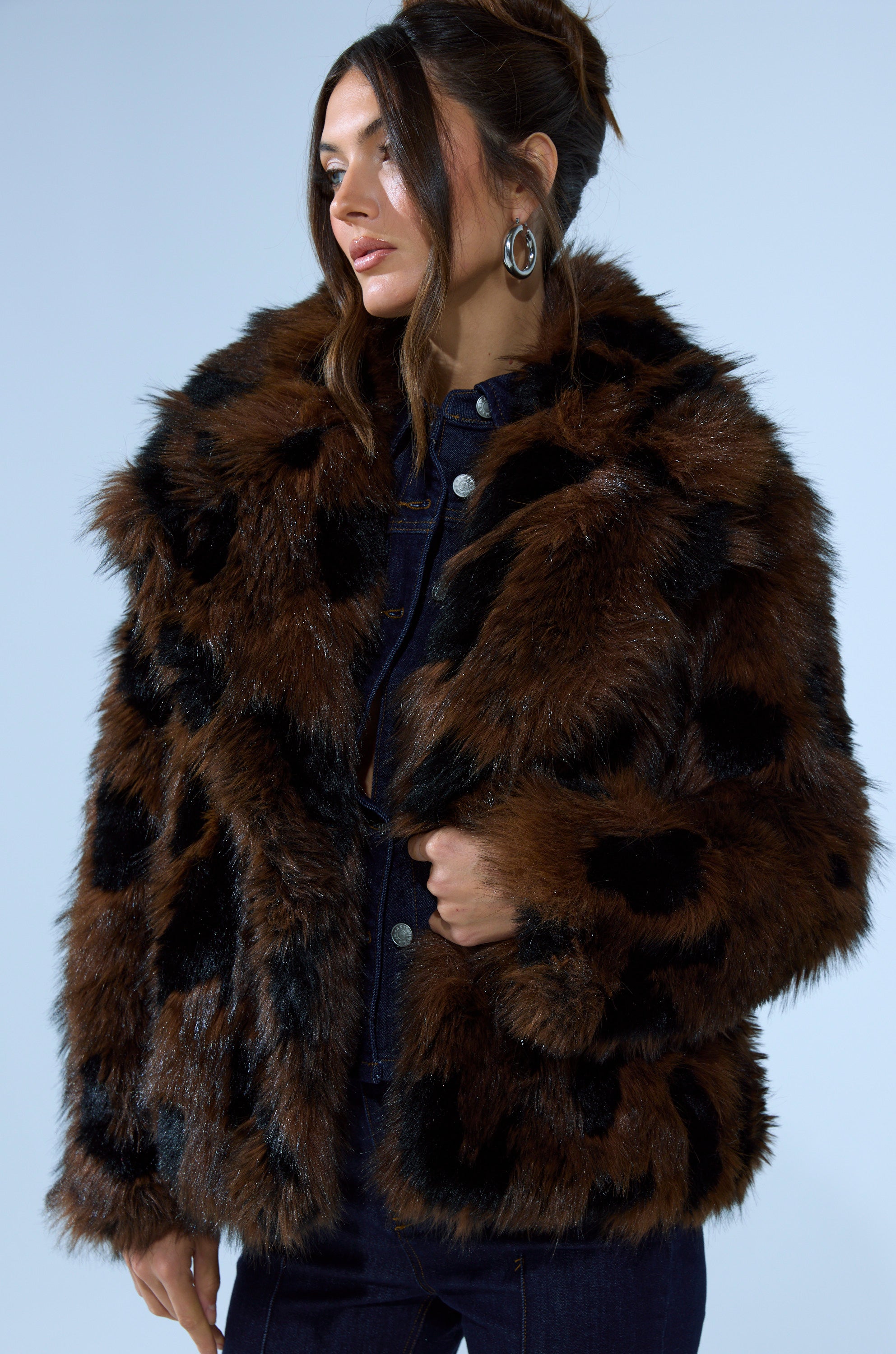 A woman wears the SHORTIE FUR COAT IN BROWN MULTI over a dark denim outfit, standing against a light blue background. Her hair is styled up and she accessorizes with large hoop earrings.