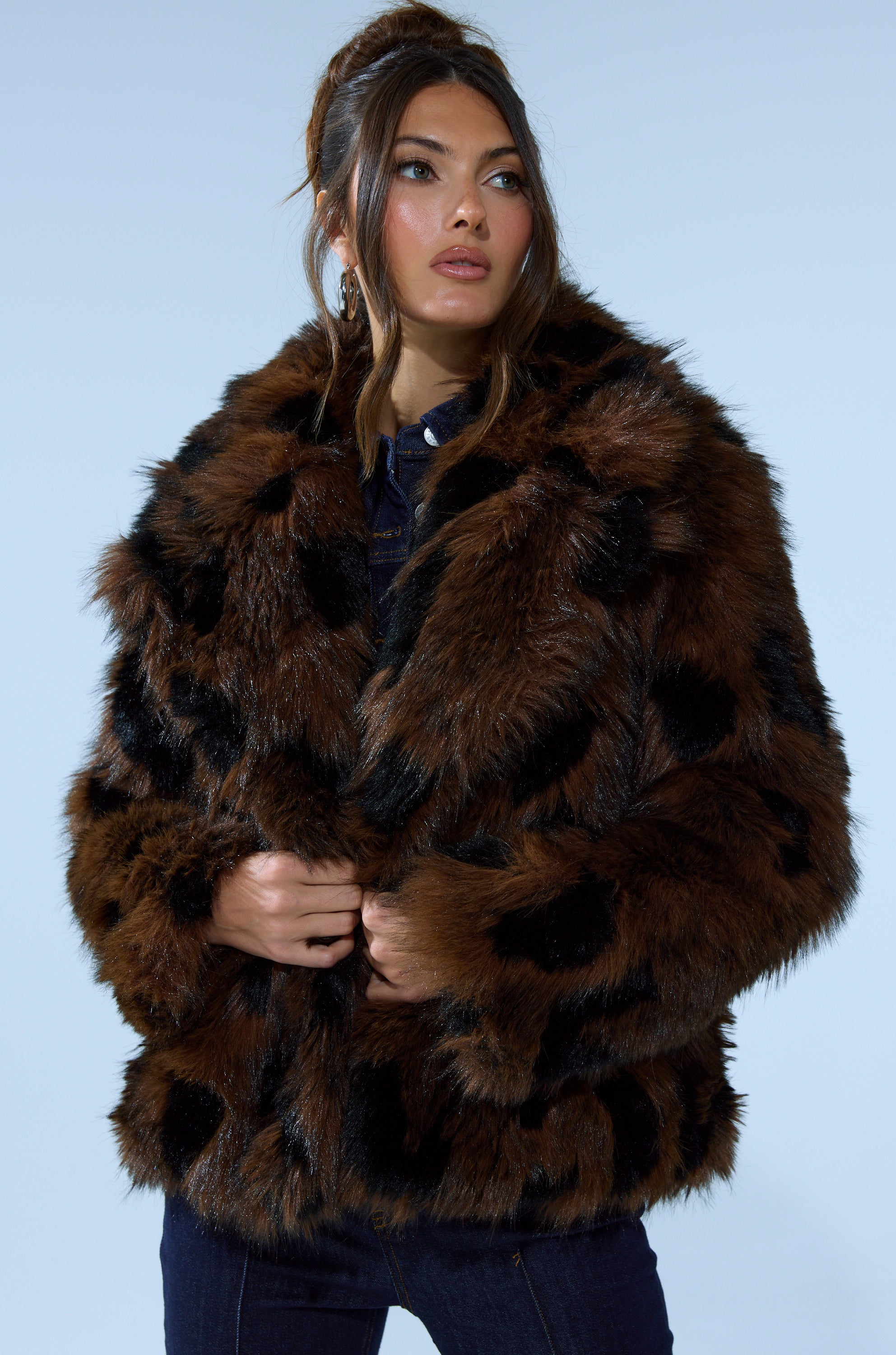 A woman stands against a light blue background, looking to the side while holding her SHORTIE FUR COAT IN BROWN MULTI closed.