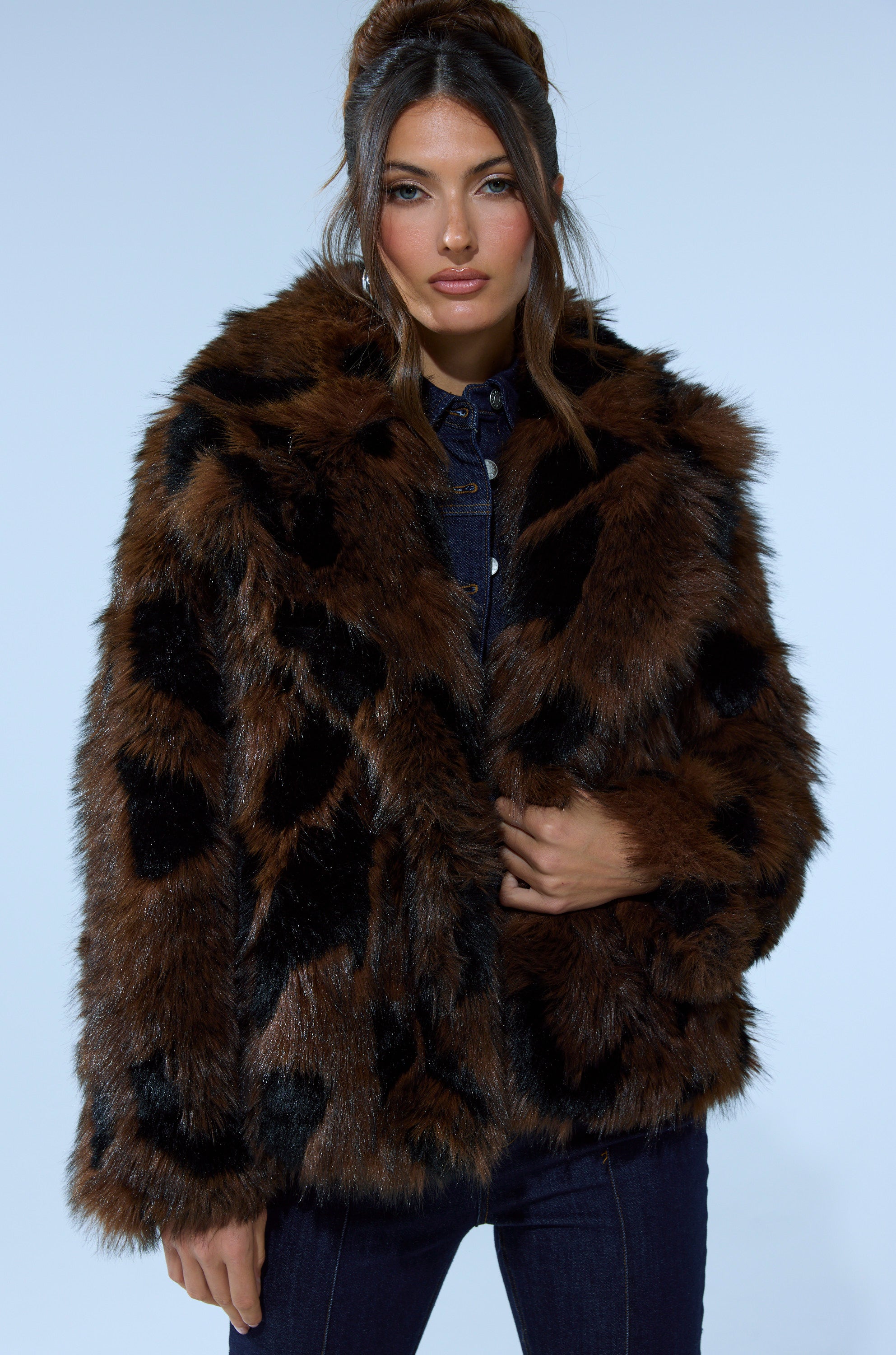 A woman with long brown hair models the SHORTIE FUR COAT IN BROWN MULTI, featuring brown and black patterns, over a dark denim outfit against a light blue background.