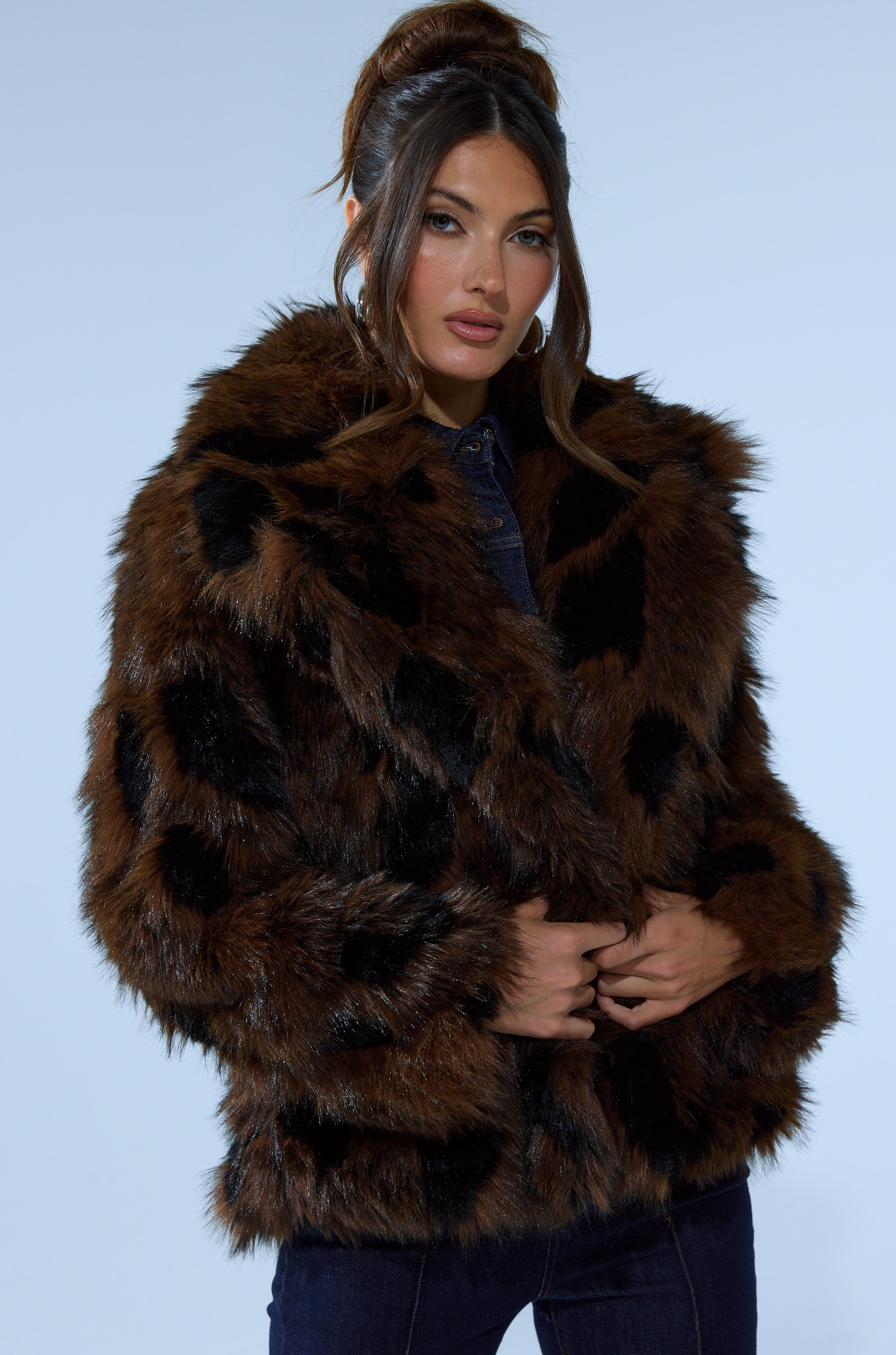 A woman with long brown hair in an updo wears the SHORTIE FUR COAT IN BROWN MULTI over a dark outfit, standing against a plain light blue background.