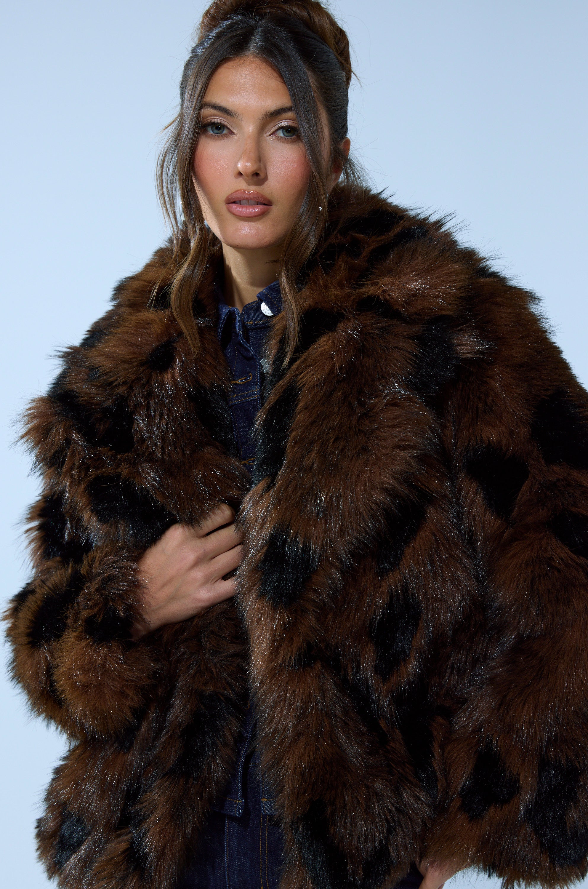 A woman with long brown hair styled in loose waves wears the SHORTIE FUR COAT IN BROWN MULTI over a dark outfit, posing against a plain light blue background.