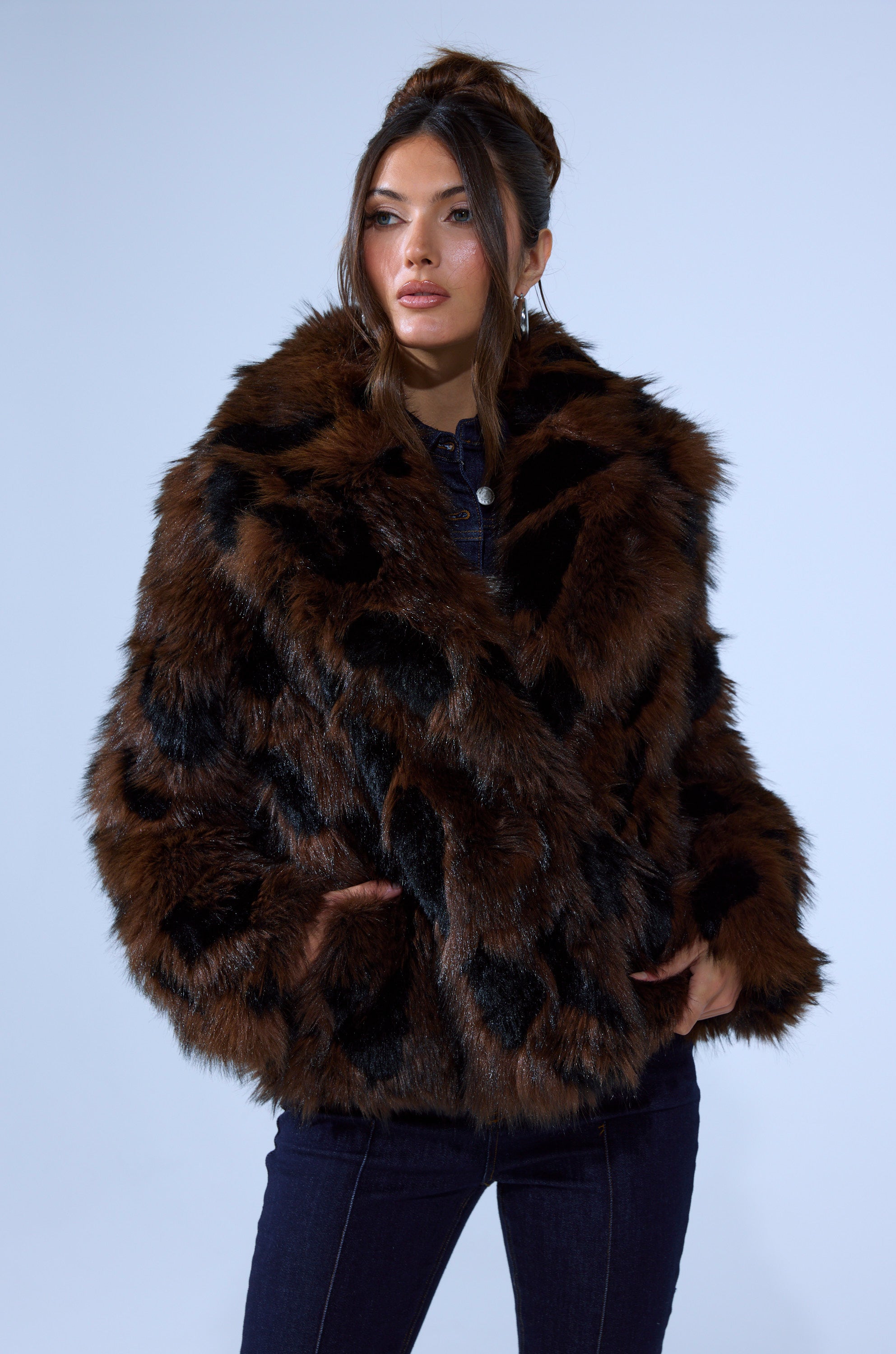 A woman with long hair styled up and dark pants wears the SHORTIE FUR COAT IN BROWN MULTI, standing with her hands in the pockets against a plain light gray background.