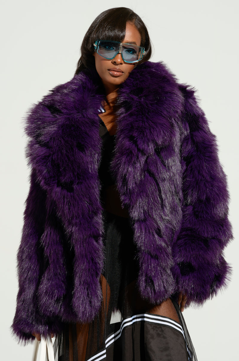 Dressed in the SHORTIE FUR COAT, an oversized faux fur piece in a striking purple shade, a person showcases bold elegance alongside trendy blue sunglasses. With long dark hair and a fashionable white bag, they stand against a simple, light-colored background.