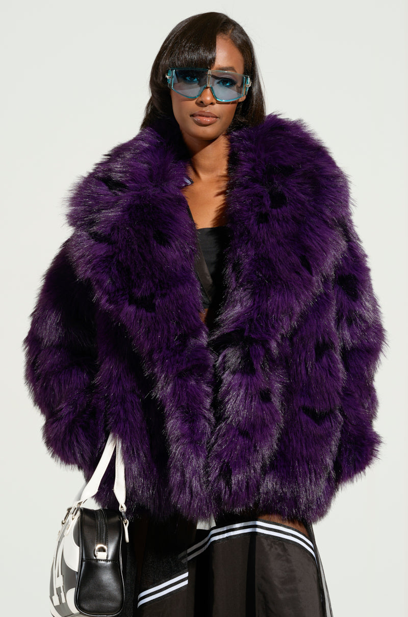 Wearing the SHORTIE FUR COAT in a striking purple hue and blue sunglasses, a person poses confidently with a black and white bag against a plain background.