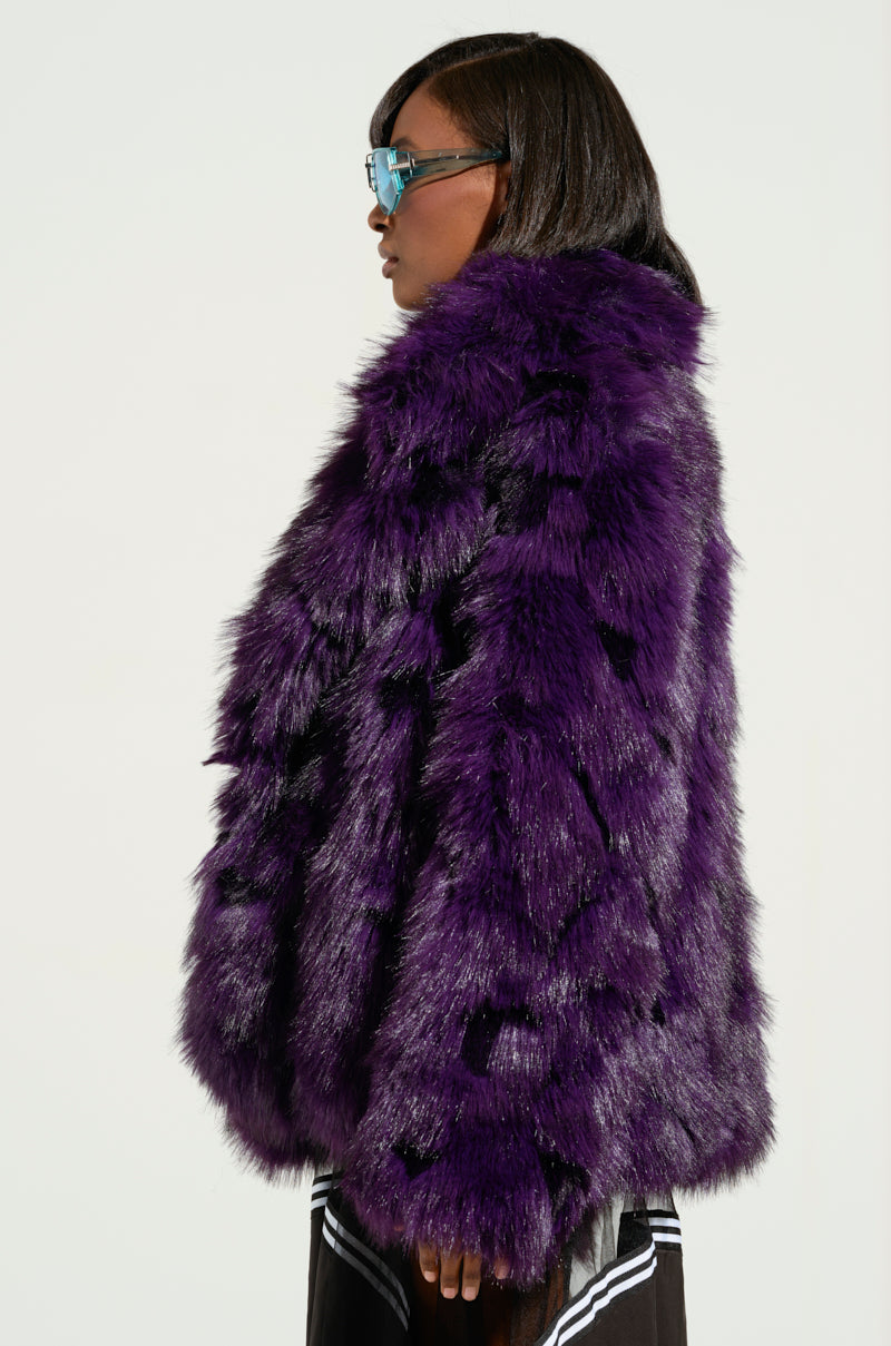 Wearing the SHORTIE FUR COAT in a vibrant purple shade with blue-tinted sunglasses, a woman stands in profile against a plain background. Her sleek, shoulder-length hair enhances her chic appearance as she looks away from the camera.