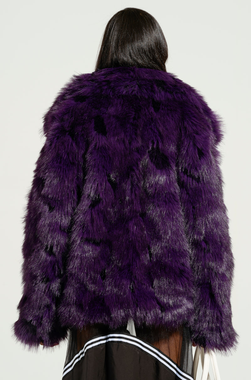 A person with long dark hair is seen from the back, wearing the SHORTIE FUR COAT in vibrant purple faux fur. The coat features a thick, plush texture and an oversized fit. They carry a white bag and wear a dark skirt with white trim, all set against a plain, light background.