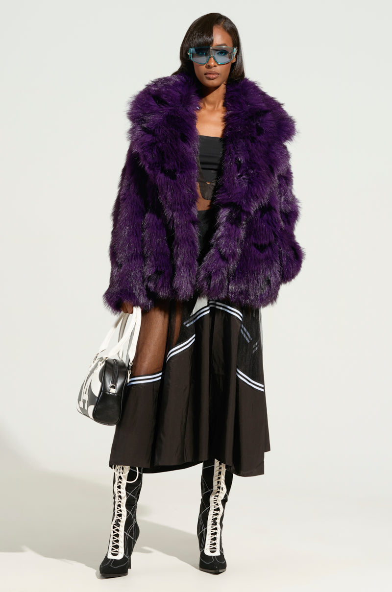 Dressed in the eye-catching SHORTIE FUR COAT, a person confidently poses in their striking purple faux fur piece. With a black outfit, chic blue sunglasses, classic white lace-up boots, and a sleek black and white handbag, they portray an audacious and stylish presence with the oversized fit of their attire.