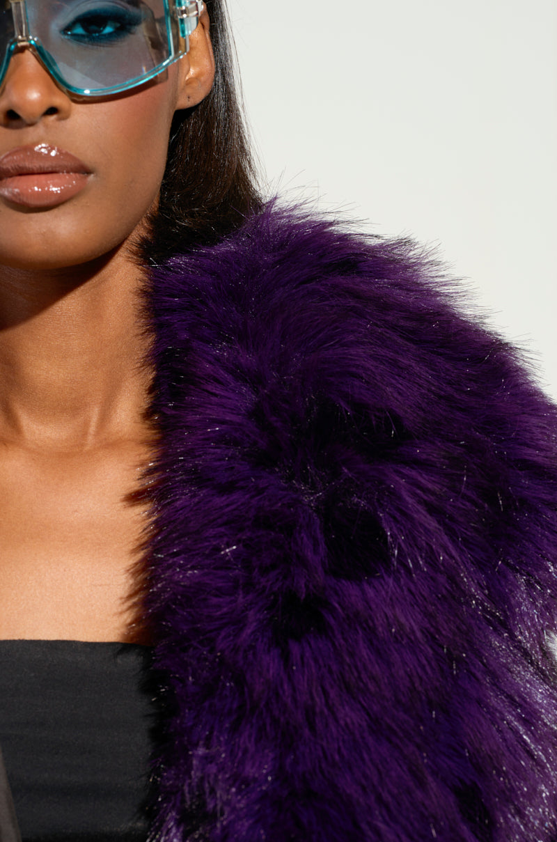 A person dressed in the SHORTIE FUR COAT, an oversized and luxurious dark purple faux fur garment, paired with blue-tinted glasses. Only the lower half of their face is visible, showcasing glossy lips against a neutral, soft beige background that enhances the coat's elegant purple tone.
