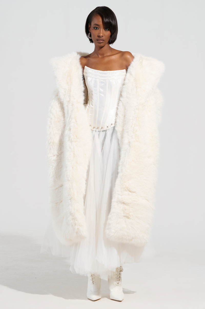 Draped in the BCARDI Long Luxe Faux Fur Trench, a person exudes elegance as the coat flows seamlessly over a strapless white corset and tulle skirt. White ankle boots complete the ensemble, with them posed gracefully against a minimalist white backdrop.