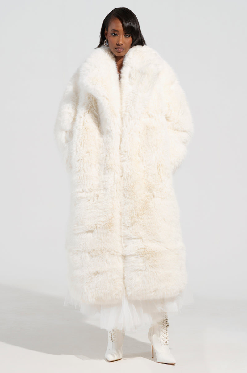 A person stands confidently in the BCARDI LONG LUXE FAUX FUR TRENCH coat from the AKIRA Label, its faux fur reaching the floor. They wear white high-heeled boots, with a white skirt peeking out from underneath against a simple light gray background.