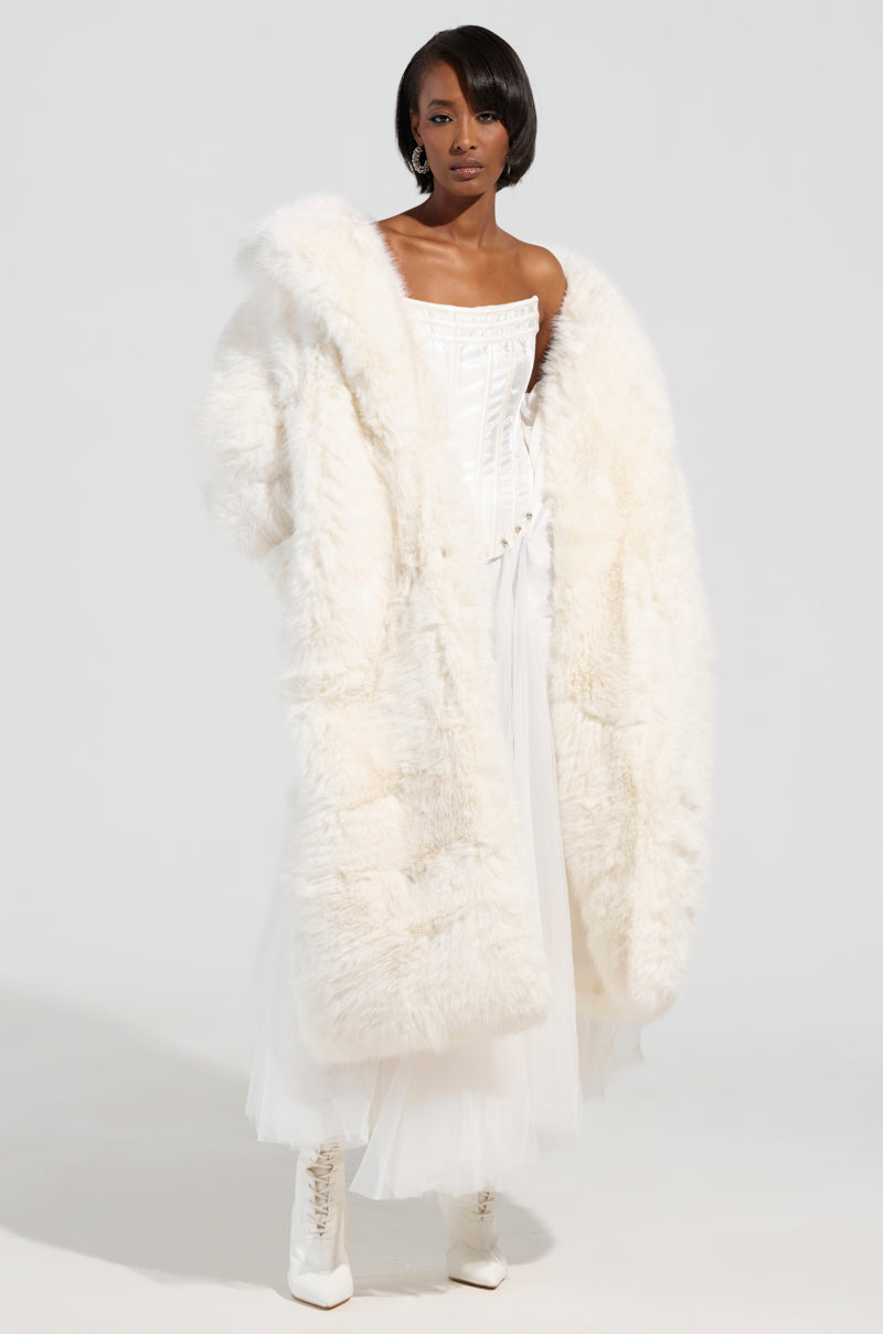 A person wearing the BCARDI LONG LUXE FAUX FUR TRENCH from the AKIRA Label over a strapless white gown with lace-up white boots stands against a plain background. The outfit blends luxurious textures with elegant styling.