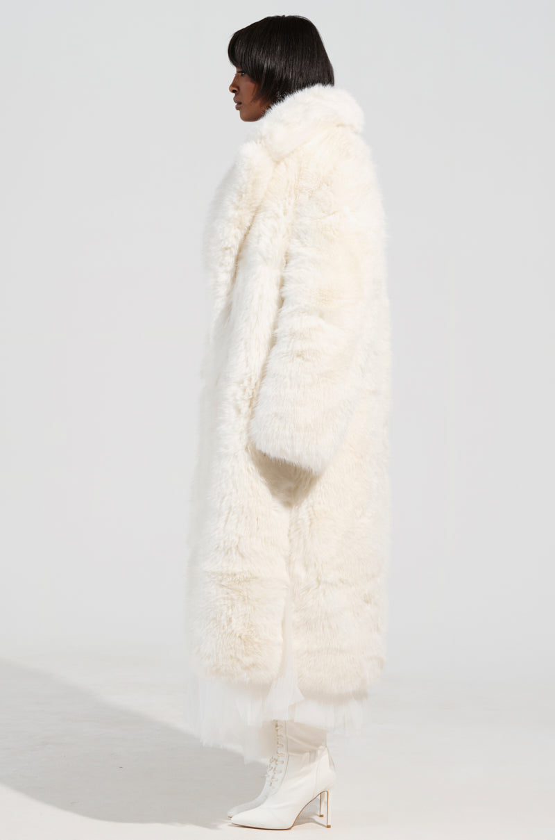 Dressed in the BCARDI LONG LUXE FAUX FUR TRENCH and high-heeled boots, an individual poses in profile against a light background. This coat from the AKIRA Label features voluminous texture and extends below the knees, offering an elegant and sophisticated appearance.