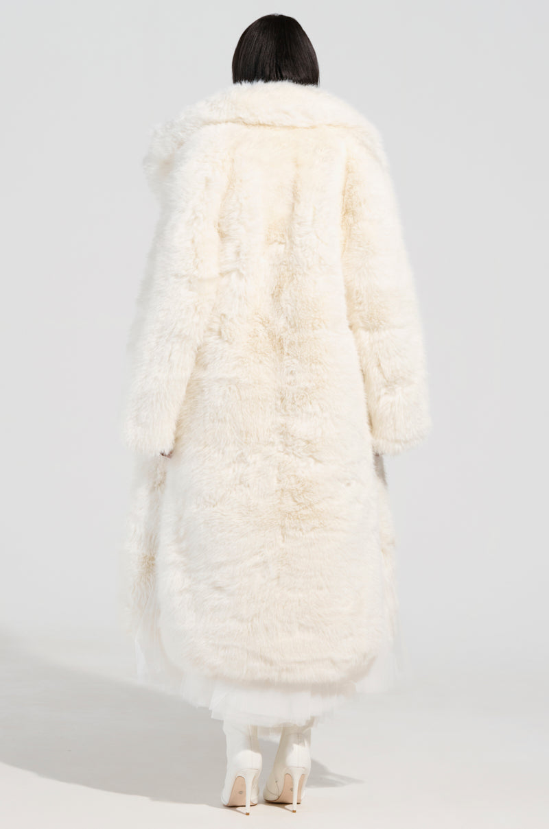 The individual is wearing the BCARDI LONG LUXE FAUX FUR TRENCH, its fluffy white design reminiscent of a luxurious trench coat. Its high collar gracefully extends down toward the ankles. Their dark hair contrasts against a light gray background as they stylishly complete the ensemble with chic white heeled boots.
