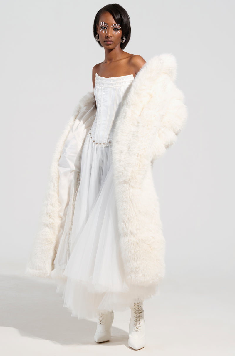 A woman stands confidently in a strapless white dress with a full skirt, draped in the BCARDI LONG LUXE FAUX FUR TRENCH over her shoulders. Stylish white boots complement her look, and ornate eye makeup features bold designs around her eyes, creating an elegant synergy between classic charm and avant-garde flair.