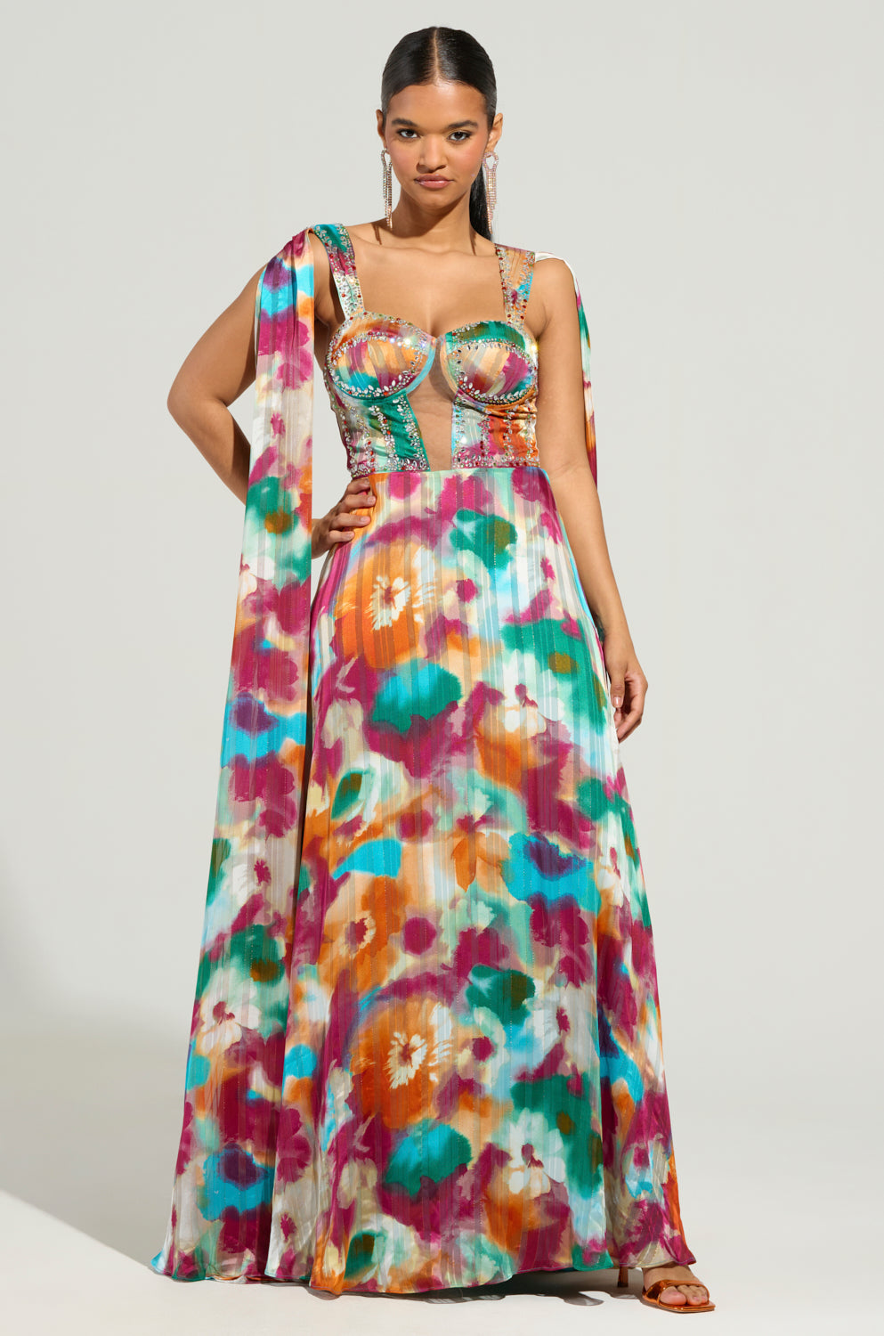 A woman wearing the ON THE RUN MAXI DRESS, adorned with a structured bodice and a long, flowing skirt in vibrant hues of pink, orange, teal, and green floral print. She stands with one hand on her hip against a neutral background.