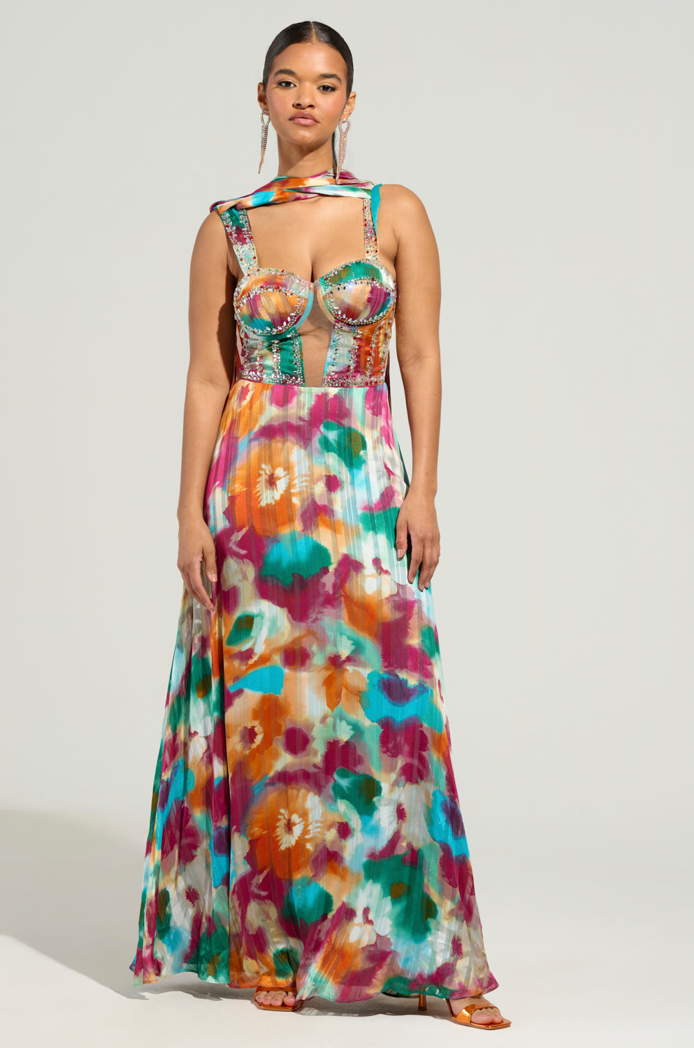 A woman stands confidently, wearing the ON THE RUN MAXI DRESS, a floral print maxi dress with a halter neck and cutout details. The vibrant patterns in shades of pink, orange, blue, and green complement her slicked-back hair as she gazes at the camera.