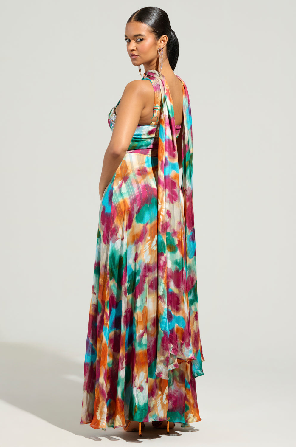 Dressed in the ON THE RUN MAXI DRESS, a vibrant chiffon gown adorned with a watercolor pattern in pinks, greens, and oranges, a person stands elegantly. The maxi dress is enhanced by a long, flowing scarf-like detail against a neutral light gray background.
