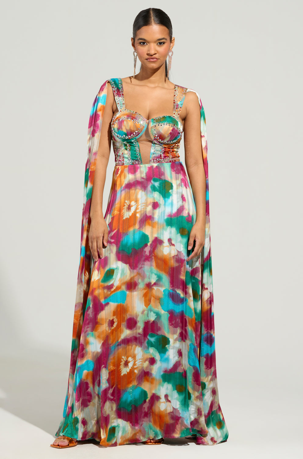 A woman in the ON THE RUN MAXI DRESS, a vibrant floral print chiffon gown, poses against a plain background. The dress showcases a structured bodice and flowing cape-like sleeves, with hues of orange, pink, green, and blue.