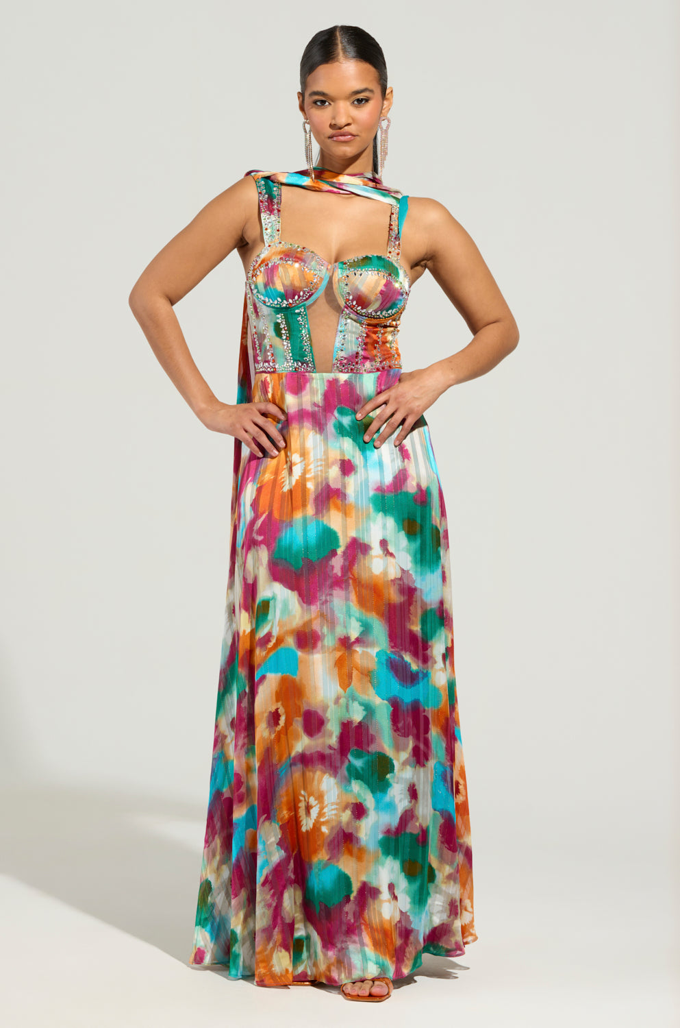 A person stands confidently wearing the ON THE RUN MAXI DRESS, a vibrant floral-print chiffon gown featuring an intricate bodice. The dress showcases multiple shades with a flowing skirt. The individual's hair is styled neatly, and they maintain a neutral expression against a plain background.
