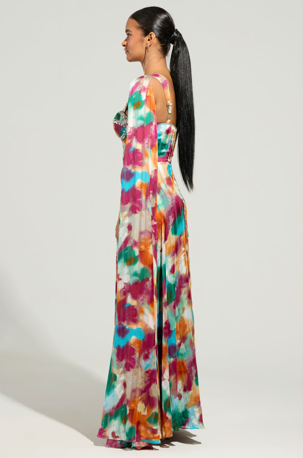 A person stands in profile, wearing the ON THE RUN MAXI DRESS, a vibrant floor-length chiffon gown with long sleeves and abstract patterns. They have long dark hair styled in a sleek ponytail, beautifully complementing the light-colored background.