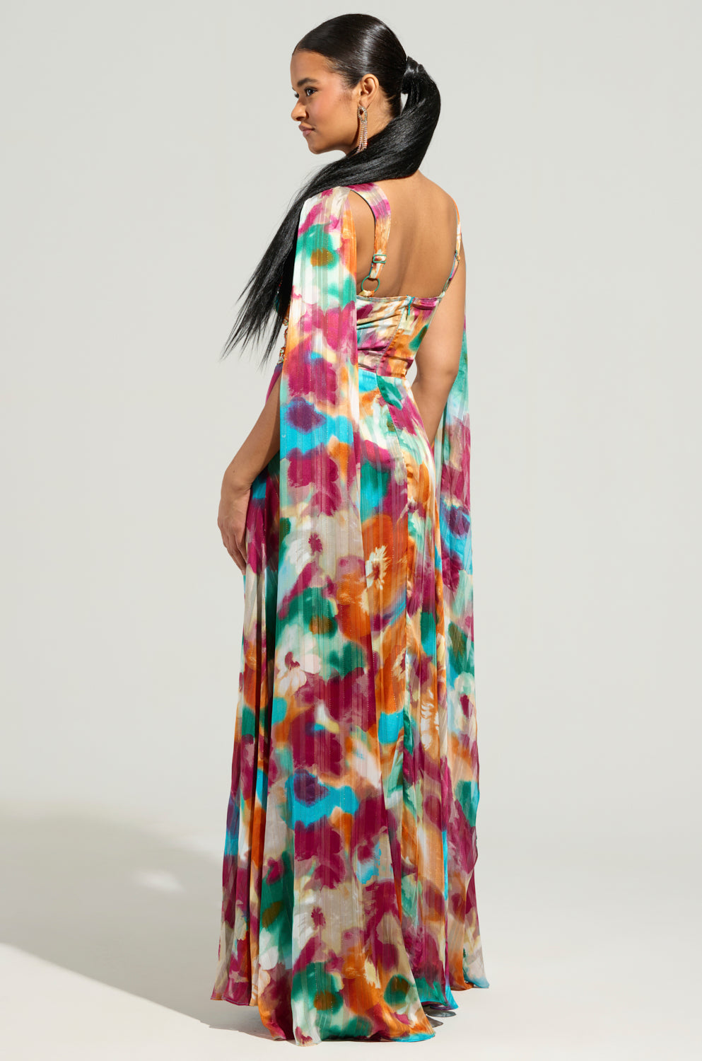 A woman stands turned slightly to the side, dressed in the ON THE RUN MAXI DRESS featuring a vibrant floral print. Her hair is styled in a sleek ponytail, and the dress resembles a colorful chiffon gown with bold splashes of red, blue, yellow, and orange.