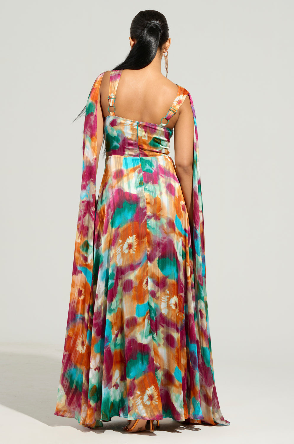 Viewing from behind, a person wears the ON THE RUN MAXI DRESS featuring a vibrant floral print. The long, flowing fabric drapes elegantly, while their hair is styled in a ponytail, creating a graceful stance against a simple backdrop.