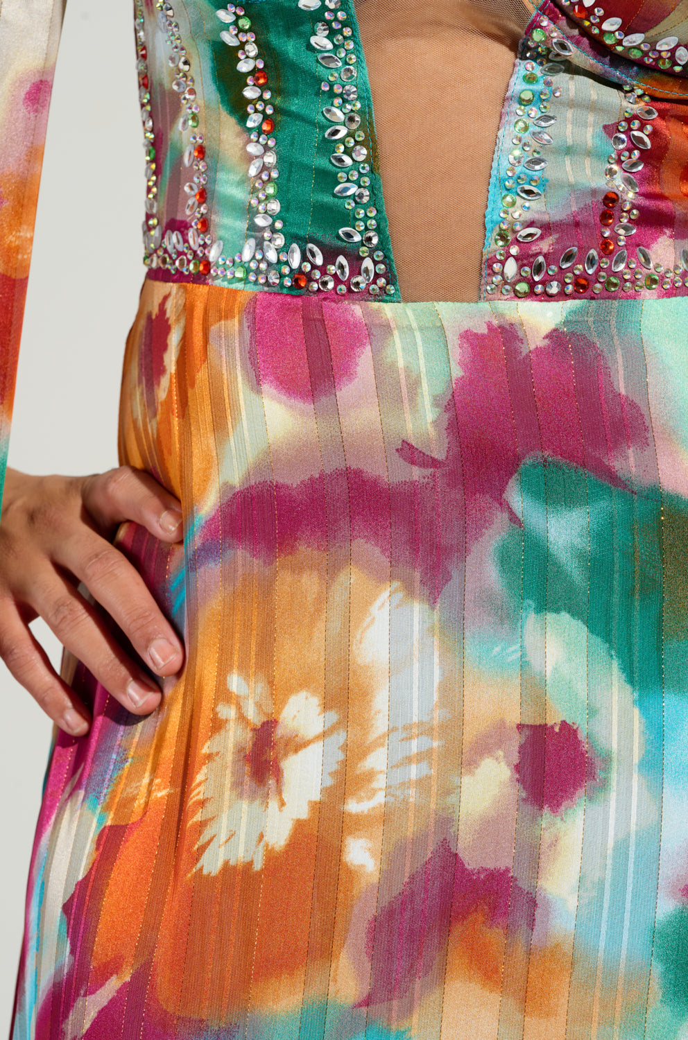 Close-up of the ON THE RUN MAXI DRESS, a vibrant chiffon gown with a floral pattern in shades of pink, orange, and green. The dress features a plunging neckline embellished with shimmering gemstones. A hand placed on the hip highlights its exquisite texture.
