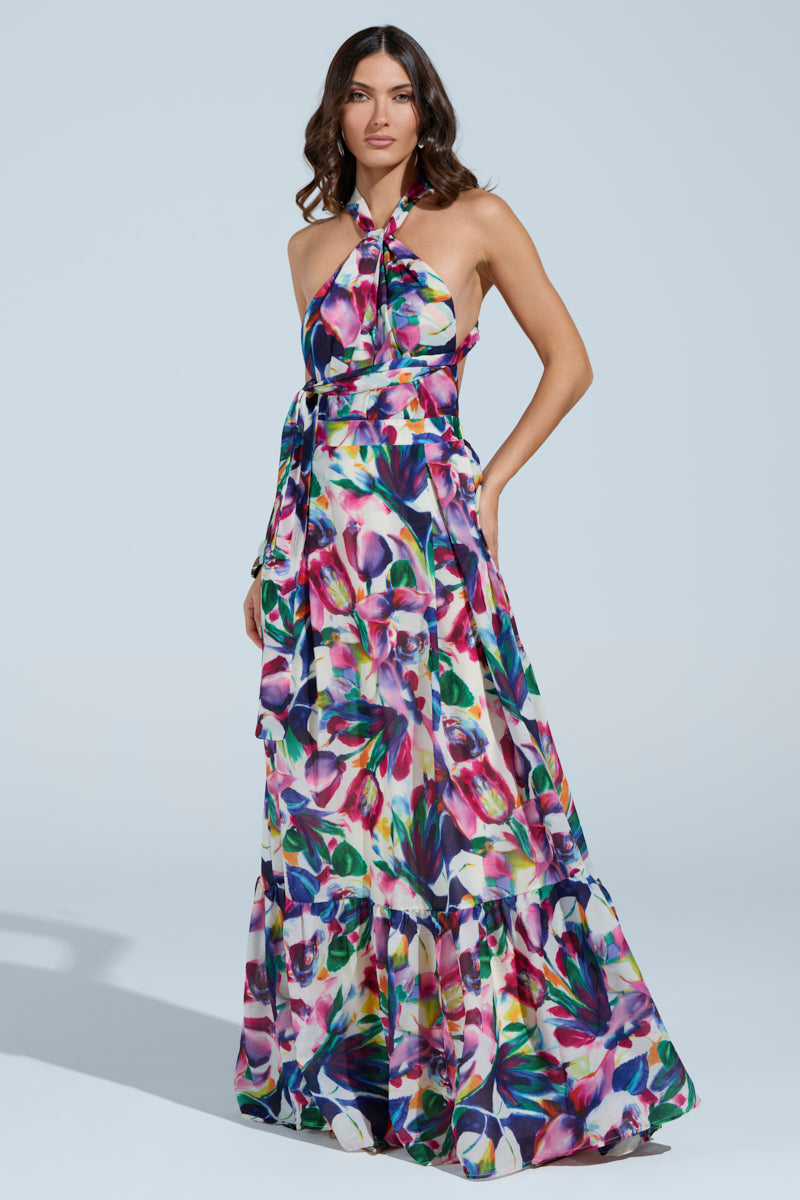 Against a light blue background, a woman poses confidently wearing the BETTER BE MAXI DRESS, featuring a vibrant floral pattern and flowing halter neckline. One hand rests on her hip, the other in her pocket, exuding elegance.