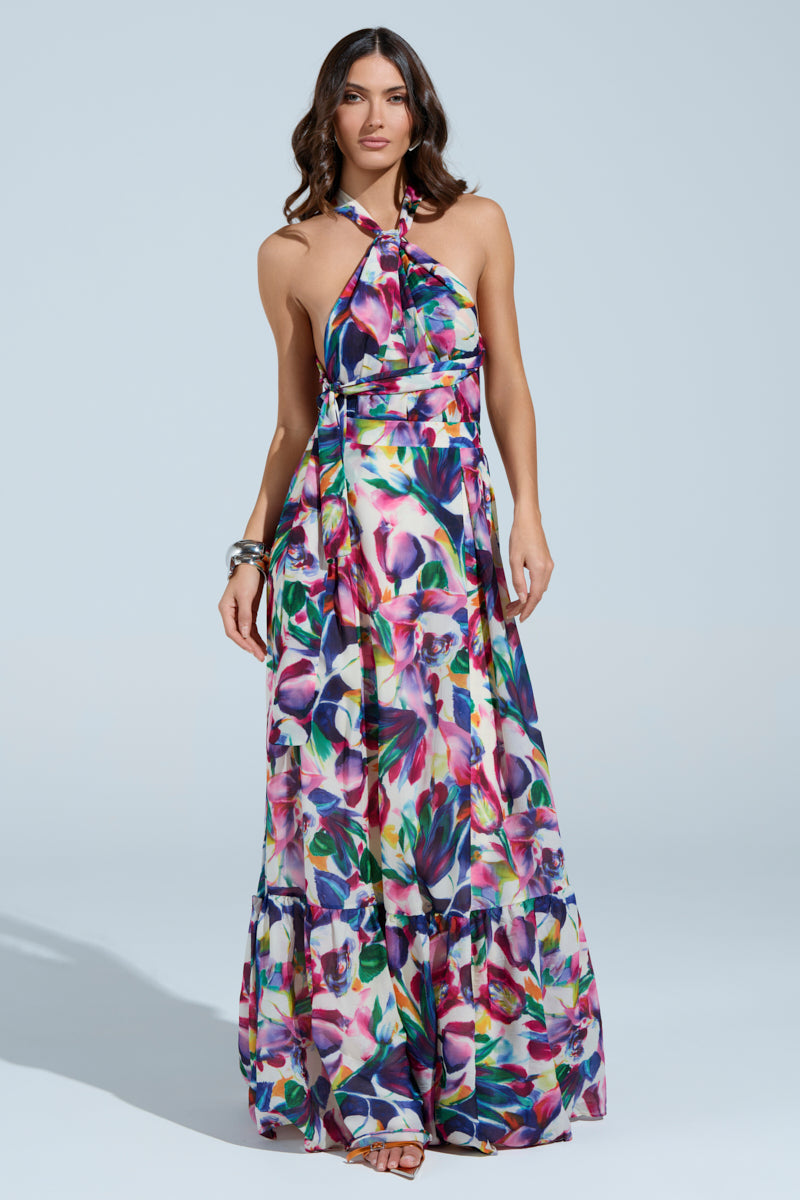A confident woman poses against a light blue backdrop in the vibrant BETTER BE MAXI DRESS, featuring a floral pattern and halter neckline. Her long, wavy hair complements the dress's elegant flow, while bracelets on her right wrist enhance her charming and poised presence.
