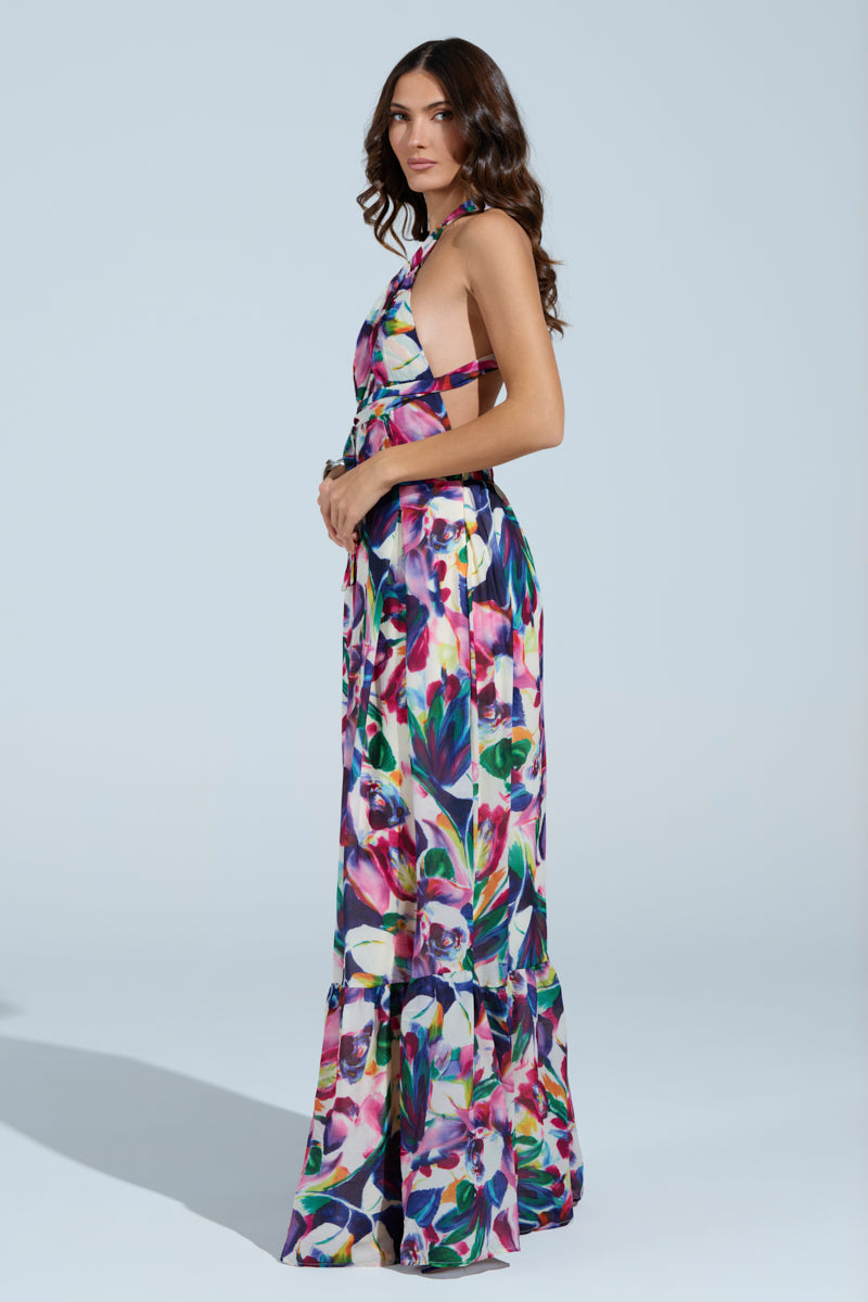 A woman in the vibrant BETTER BE MAXI DRESS with a halter neckline stands against a plain background. Her long brown hair cascades over her shoulders in loose waves as she poses sideways, hands gently resting in front.