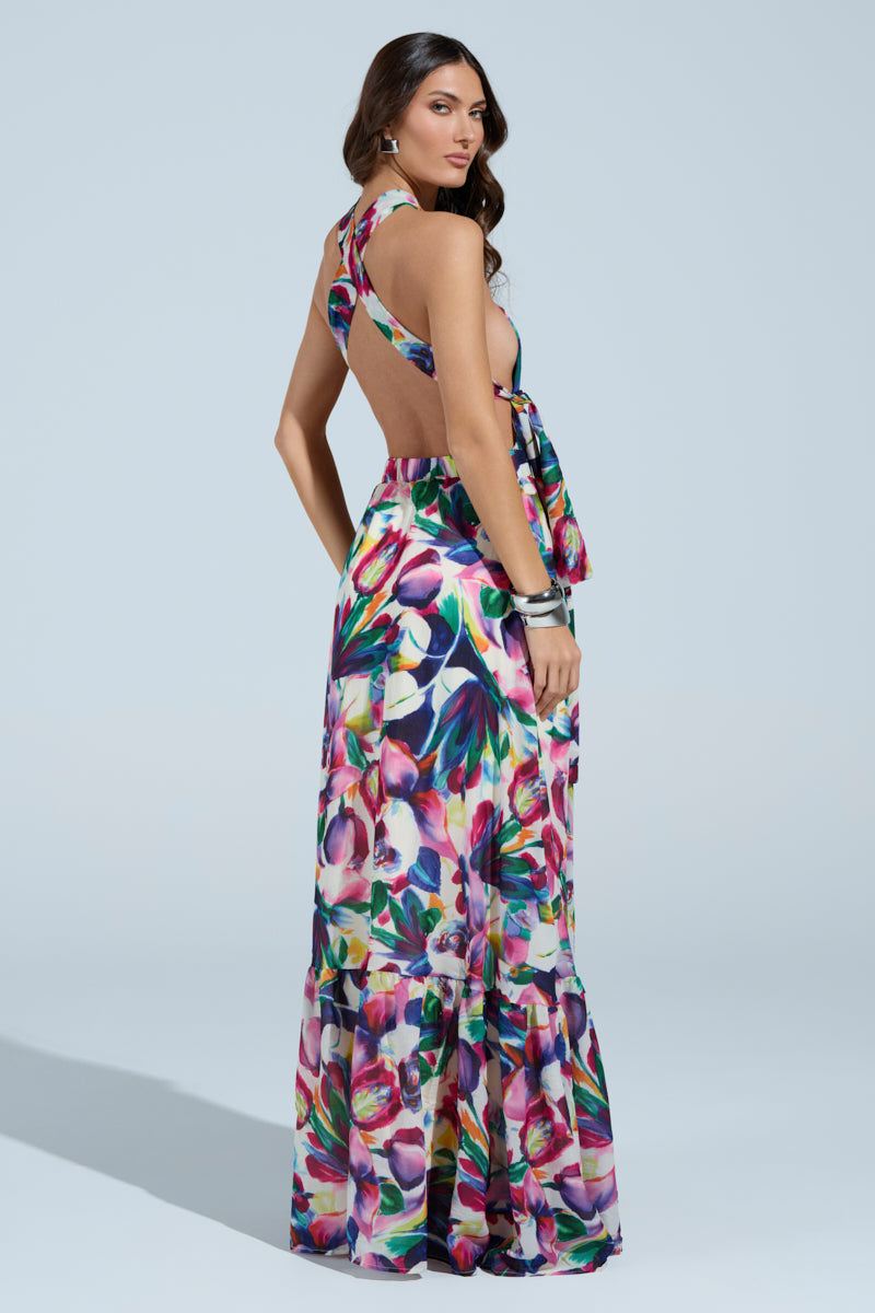 Against a light blue background, a woman models the BETTER BE MAXI DRESS, featuring a floral pattern with a halter neckline and open back. Her long dark hair cascades over her shoulders, and she wears a silver bracelet. She poses relaxedly, glancing over her shoulder.