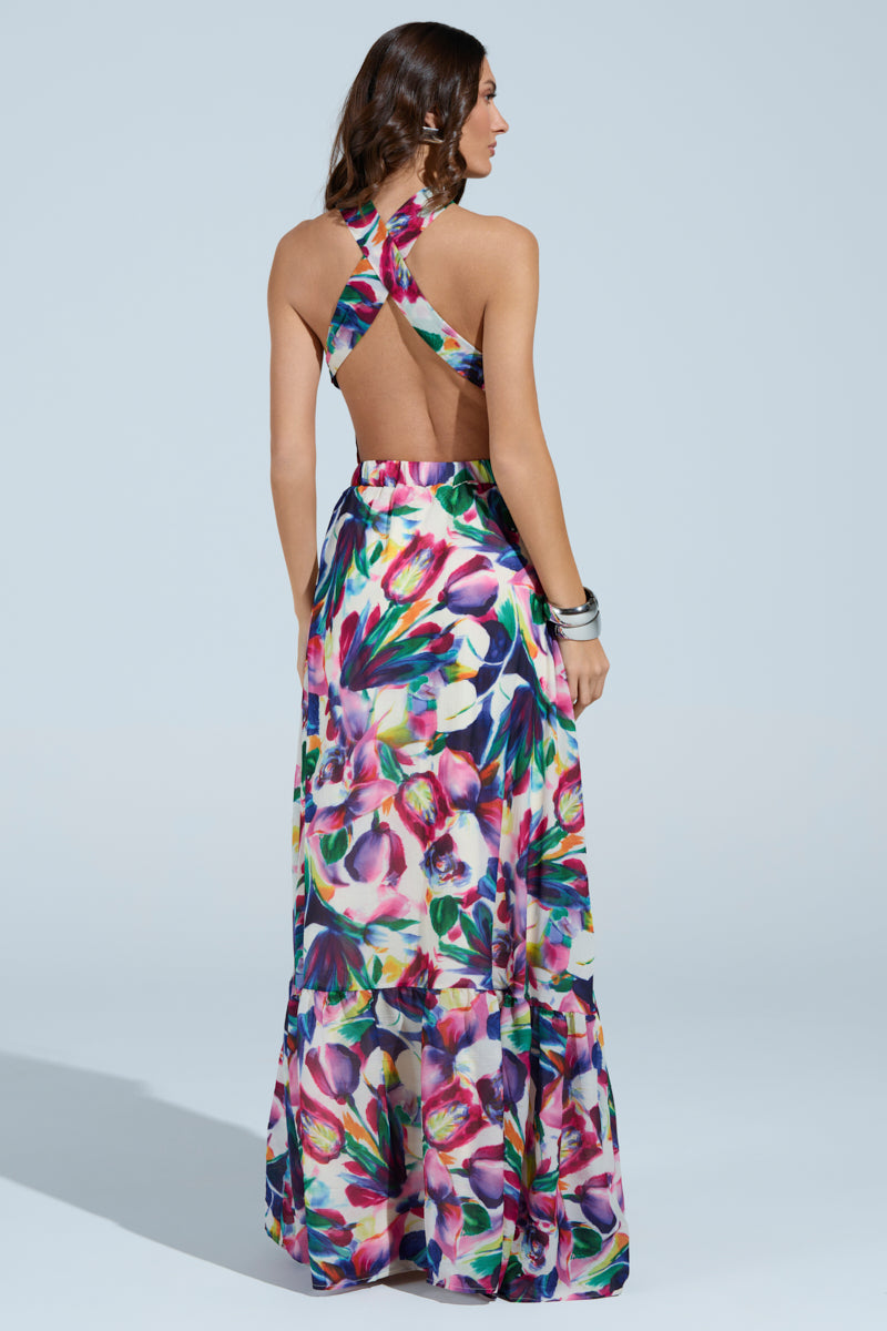 A woman models the BETTER BE MAXI DRESS, showcasing its vibrant floral pattern, crisscross open back, and halter neckline. She stands against a light blue backdrop with her long wavy hair and bracelets accenting the dress's full length as she gazes away from view.