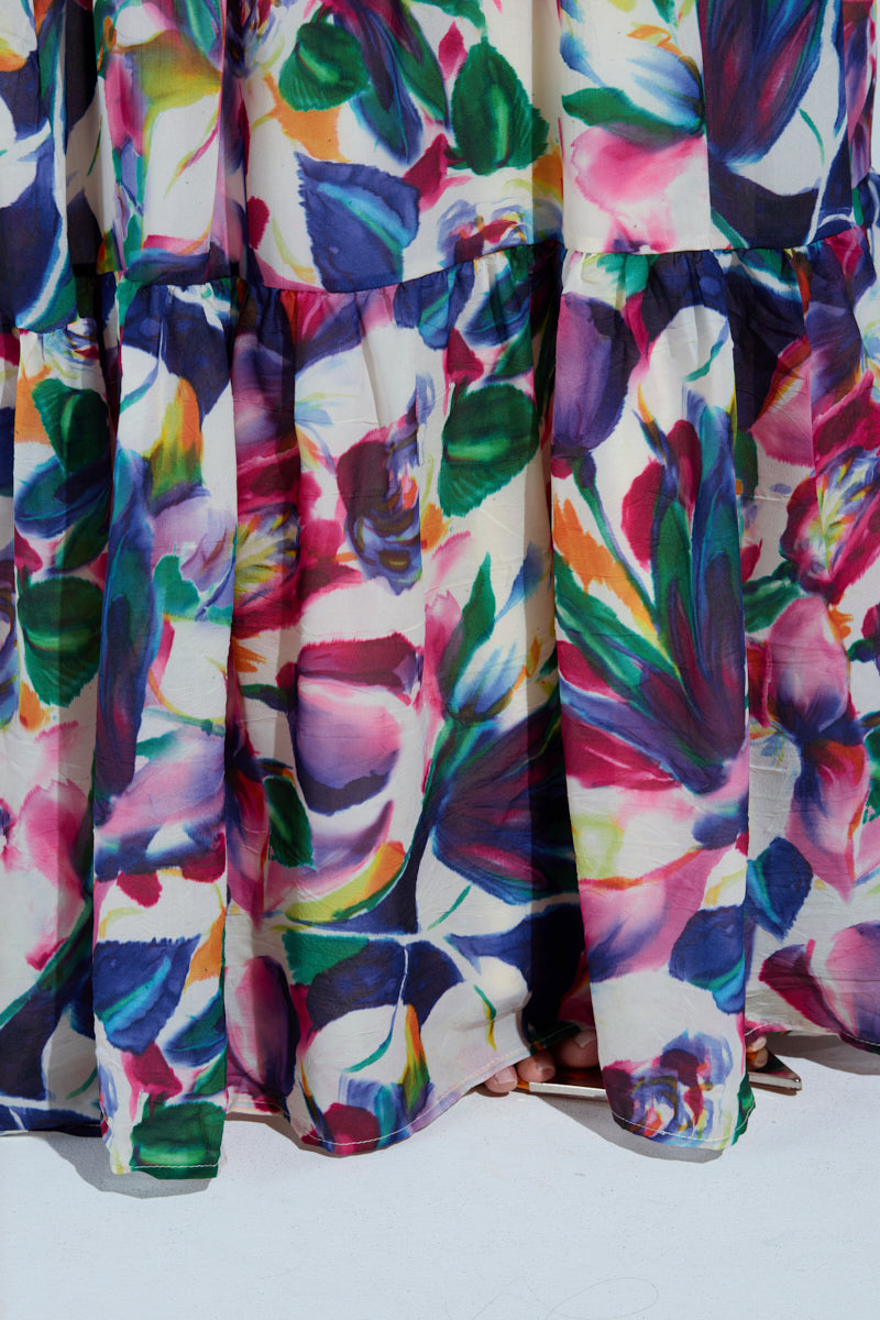 The BETTER BE MAXI DRESS, featuring a vibrant floral pattern in blues, greens, pinks, and purples on white, gracefully drapes over feet in strappy high-heeled sandals. Its halter neckline adds an elegant touch.