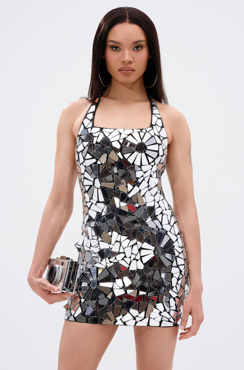 A woman stands facing forward in the ALL EYES ON ME MIRRORED MINI DRESS, its body-hugging silhouette covered in geometric mirror pieces. She holds a metallic clutch, wears hoop earrings, and has straight dark hair against a plain white background.