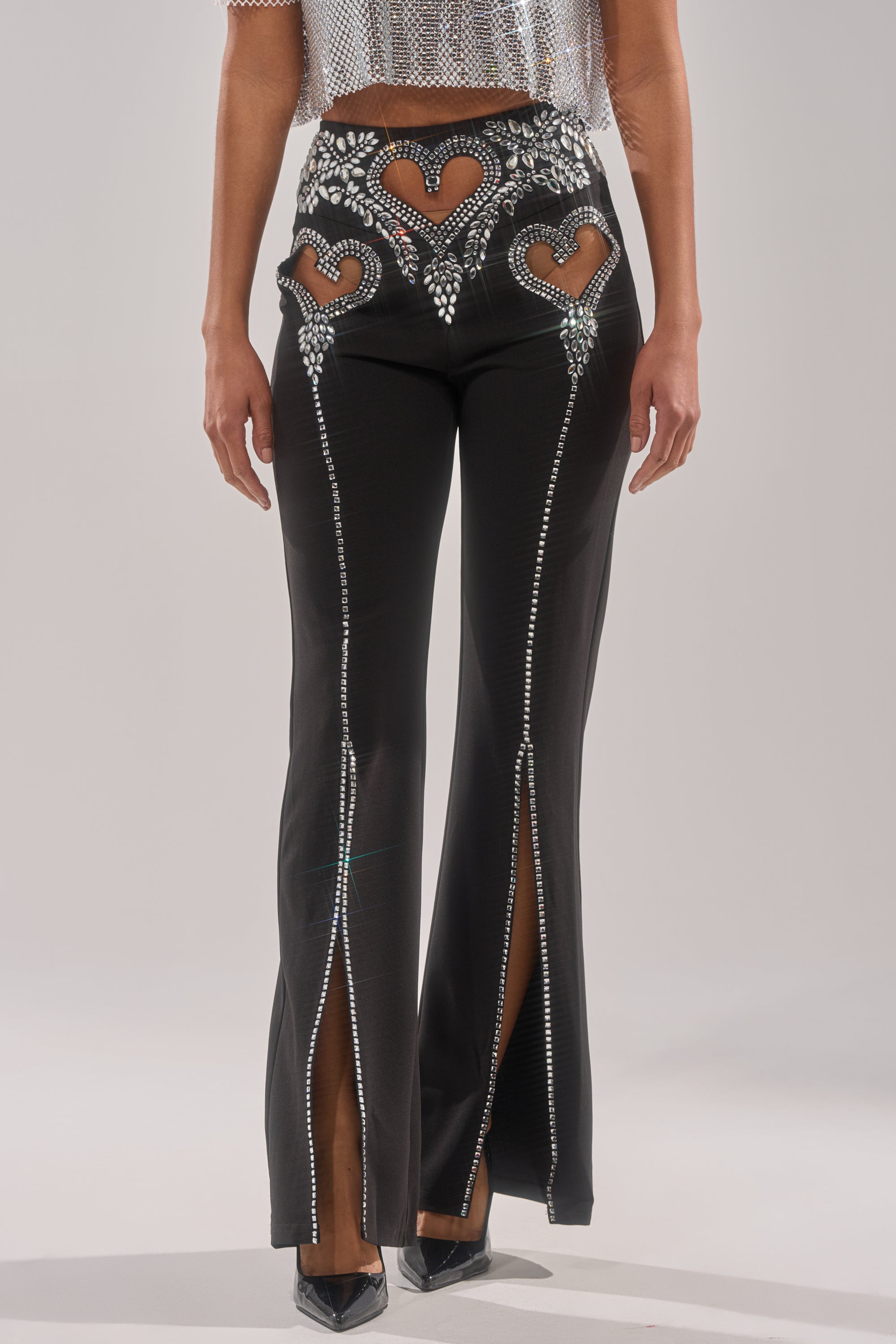 A person wears the LOVE FOR YOU RHINESTONE TROUSER—black flared pants with heart cutouts, crystal embellishments, front slits revealing high heels—paired with a sparkly cropped top, all set against a light background.