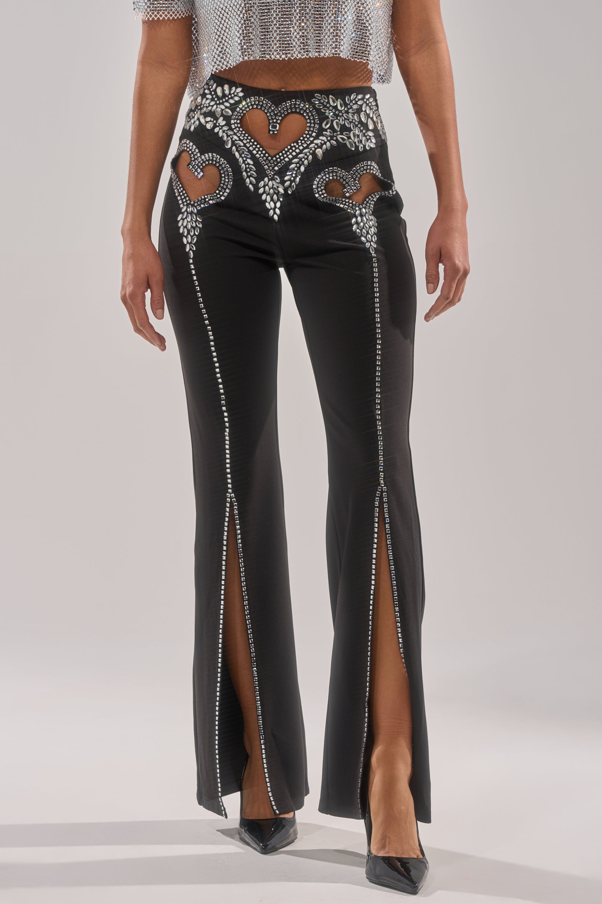 A woman wears the LOVE FOR YOU RHINESTONE TROUSER featuring black heart cutouts, rhinestone embellishments, flared legs, and ankle slits. She pairs them with a silver mesh crop top and black heels for a dazzling look.