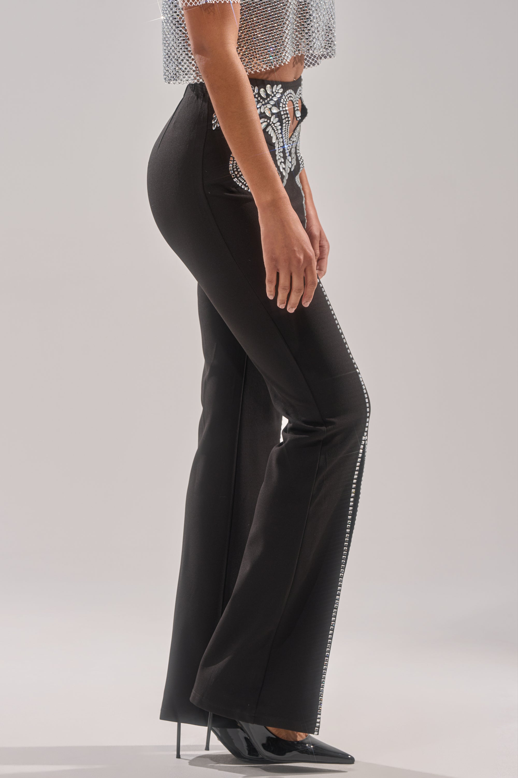A person stands sideways against a plain background wearing the LOVE FOR YOU RHINESTONE TROUSER, featuring black flared legs with rhinestone details, paired with a silver sequined crop top and black pointed-toe heels.