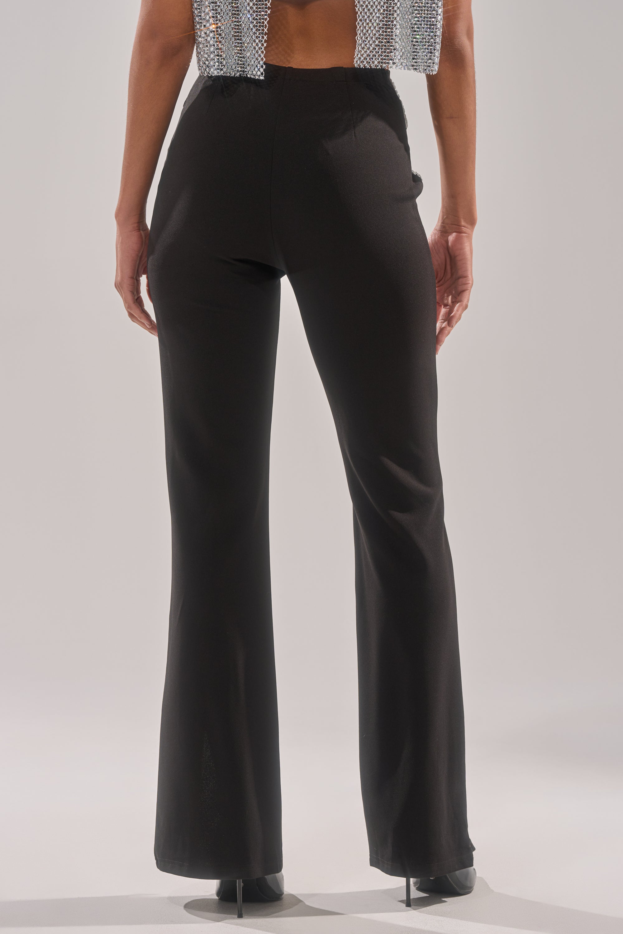 A person stands with their back to the camera in LOVE FOR YOU RHINESTONE TROUSER—sleek black flared pants—paired with a sparkly silver top, against a plain light background.