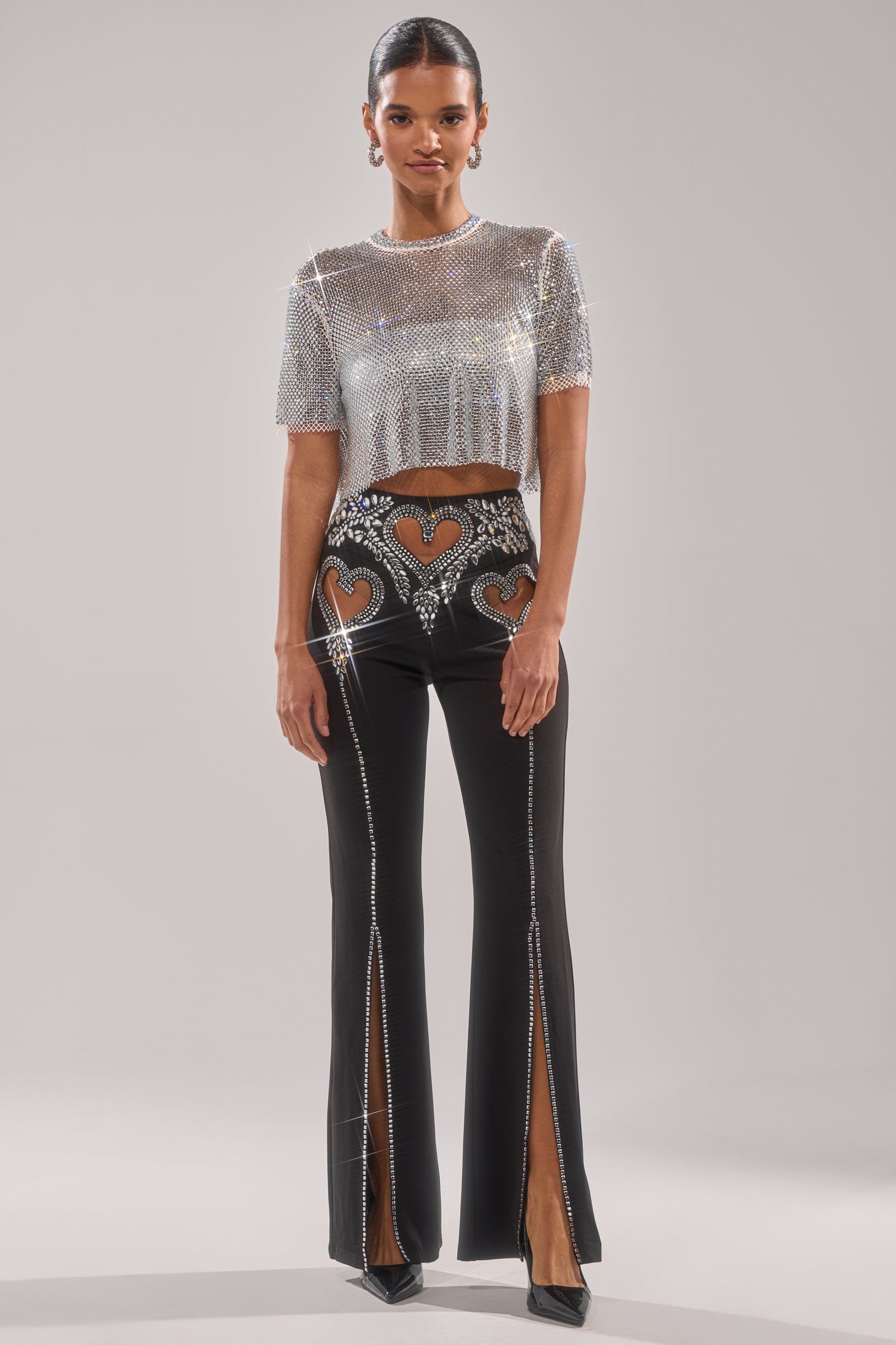 A woman wears the LOVE FOR YOU RHINESTONE TROUSER—black flared-leg pants with heart cutouts, silver rhinestone embellishments, and front slits—paired with a silver sequin crop top and pointed black heels.