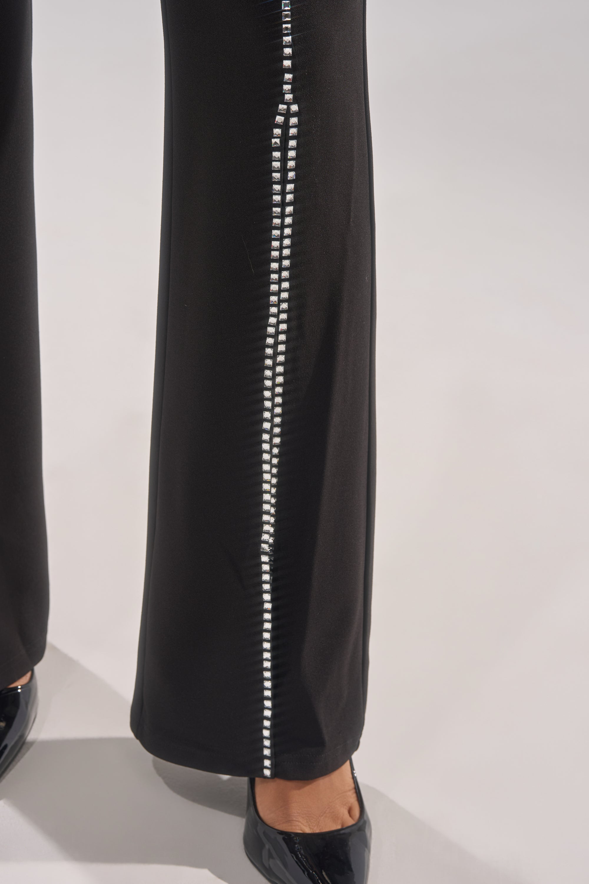 A close-up of the lower legs shows a person wearing LOVE FOR YOU RHINESTONE TROUSER with black flared legs, a vertical white decorative stripe, and black patent shoes against a plain light background.