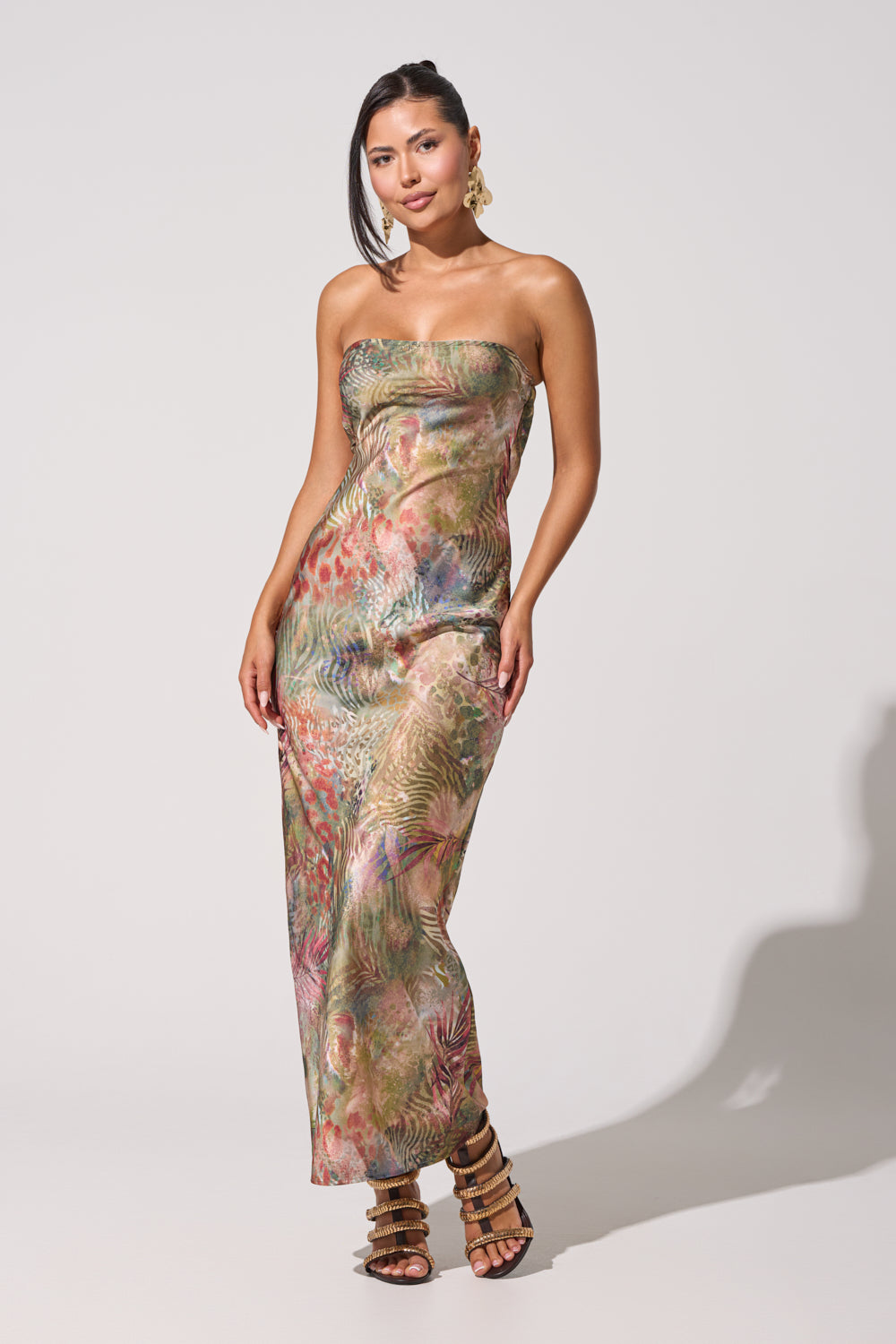 A woman poses in the IT GIRL SATIN MAXI DRESS, featuring a strapless, vibrant tropical print. She accessorizes with large hoop earrings and strappy high-heeled sandals, her straight hair loose against a plain white background. 1