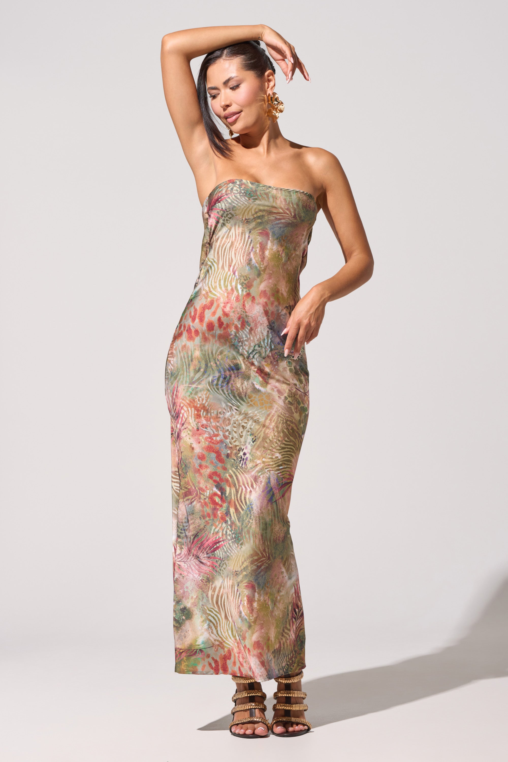 A woman poses in the IT GIRL SATIN MAXI DRESS, a strapless satin maxi with a colorful abstract animal print. She pairs it with strappy high-heeled sandals and large earrings, one arm raised above her head against a plain background. 0