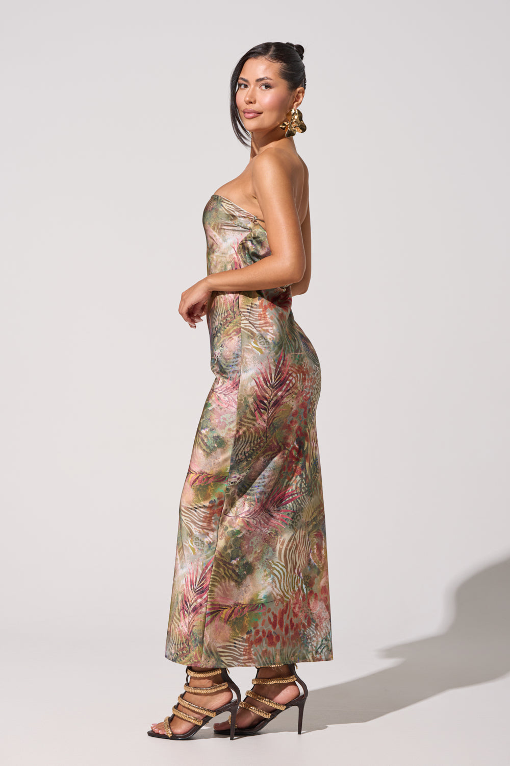 A woman stands sideways in the IT GIRL SATIN MAXI DRESS, featuring a strapless tropical print, paired with gold earrings and strappy heels against a plain light background. 3
